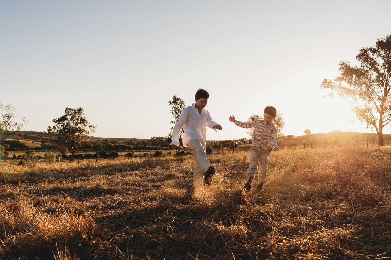 Two people in white outfits run joyfully through a sun-drenched field at golden hour.