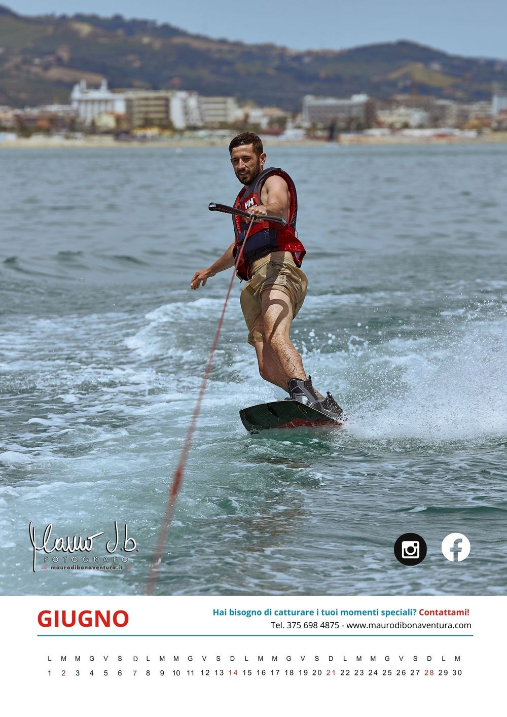 A wakeboarder rides across the water with mountains and buildings visible along the coastline in the background.