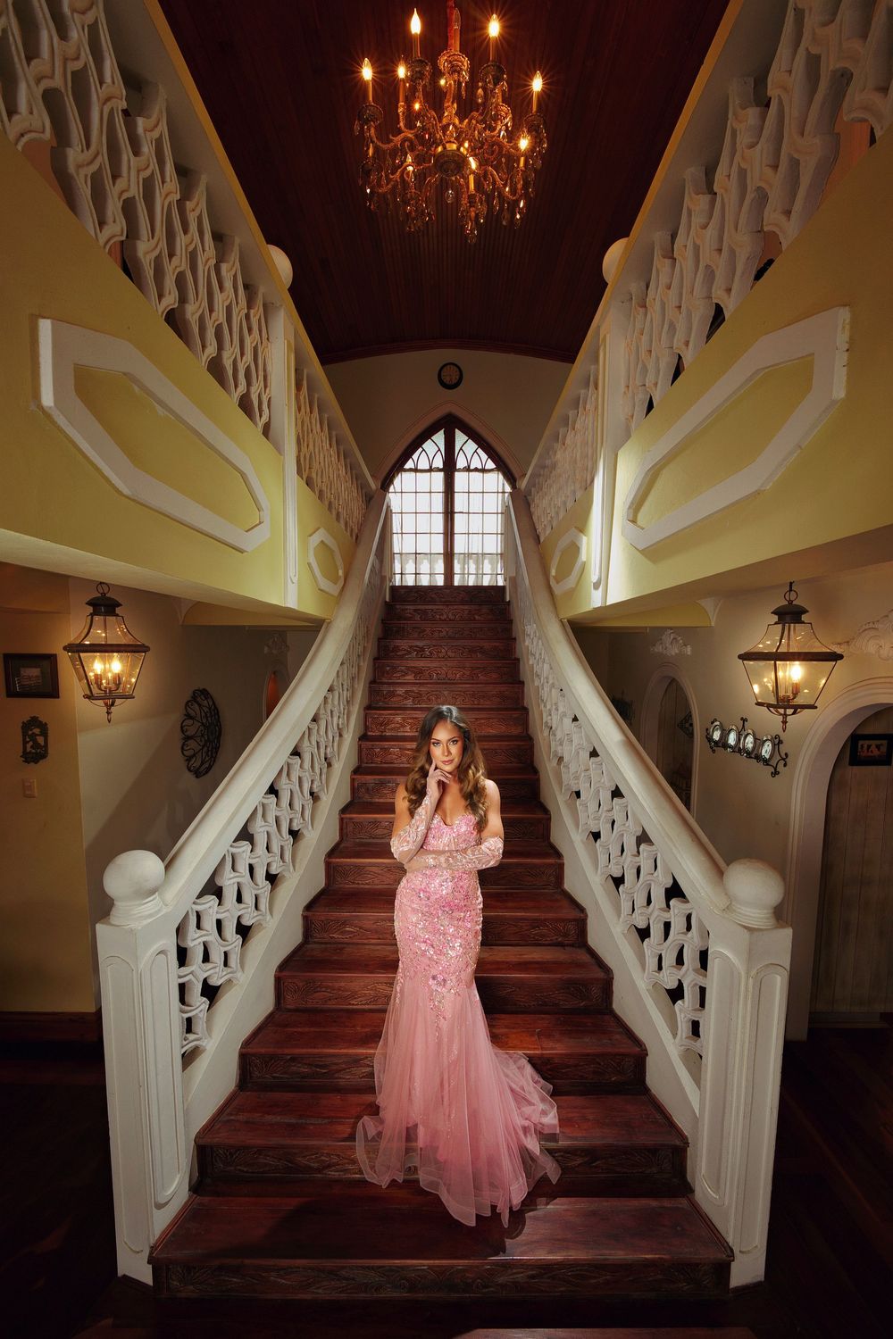 Elegant pink ball gown photographed on a grand wooden staircase with ornate white railings and chandeliers.