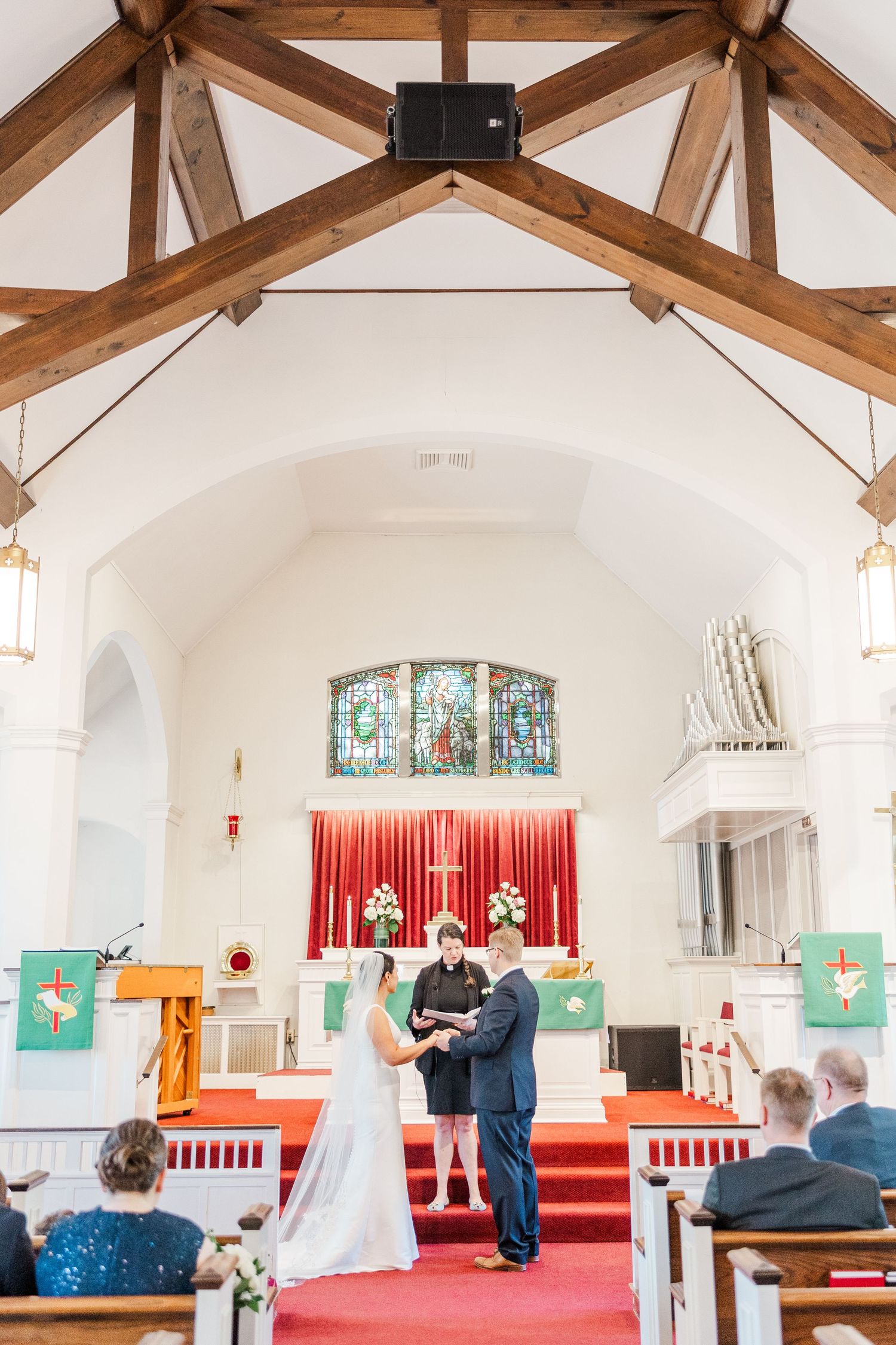Interior of a church with exposed wooden beams, stained glass windows and red carpet during a wedding ceremony.