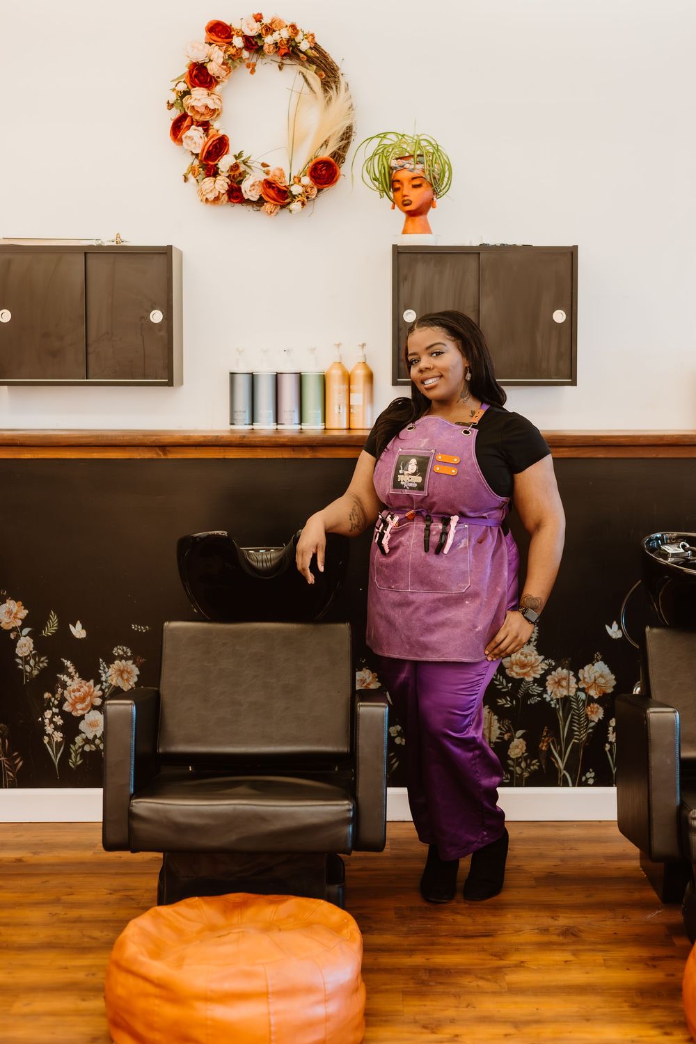 A person in a purple shirt sits and poses playfully on dark wooden chairs in a room decorated with autumn wreaths.
