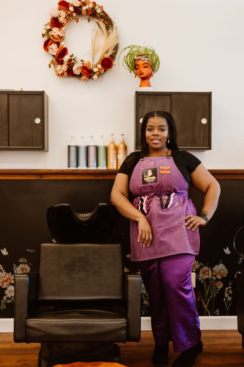 Hair salon professional wearing purple apron and pants stands in front of wall-mounted storage and decorative wreath.