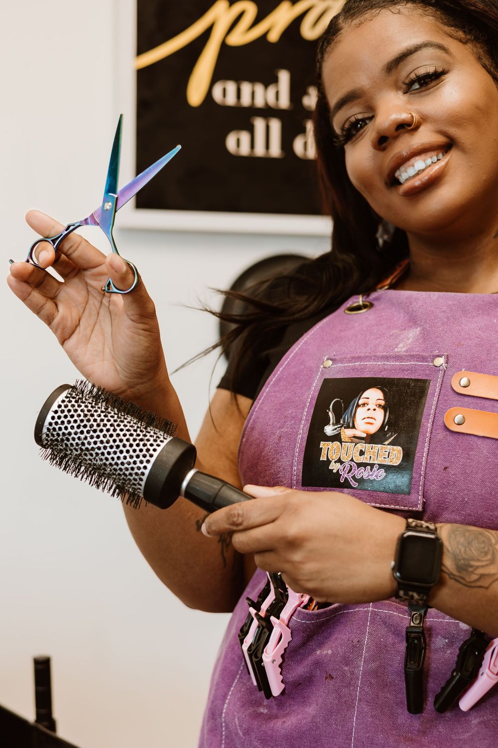 Professional hairstylist holds styling tools while wearing a purple apron in a salon setting.