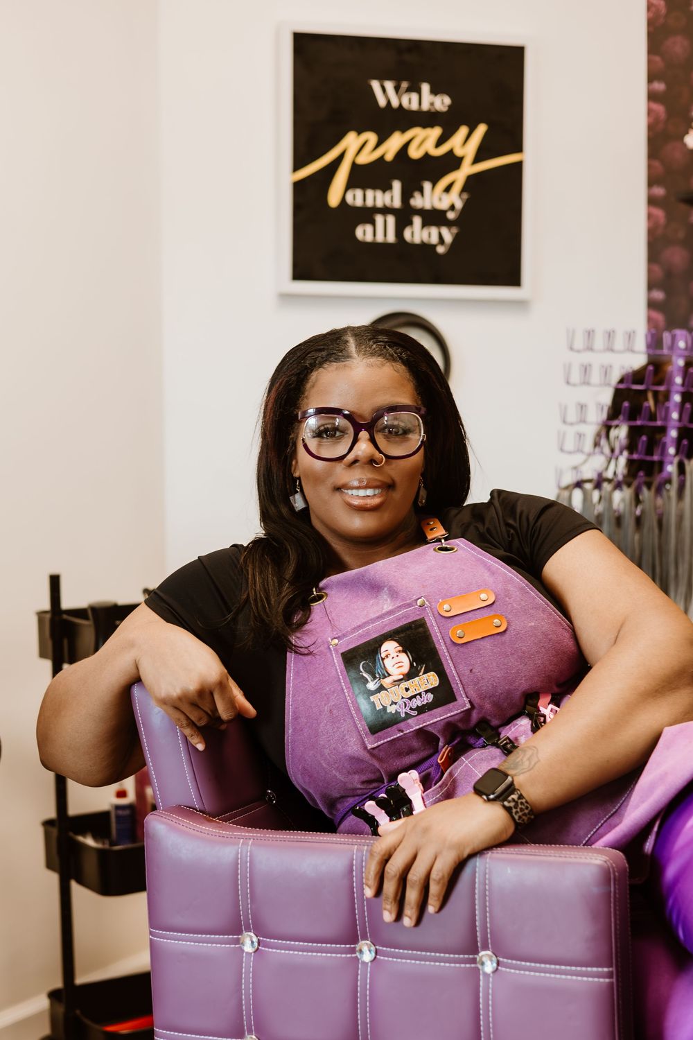 A person sits on a purple leather chair beneath a 'Wake pray and slay all day' sign in a stylish interior setting.