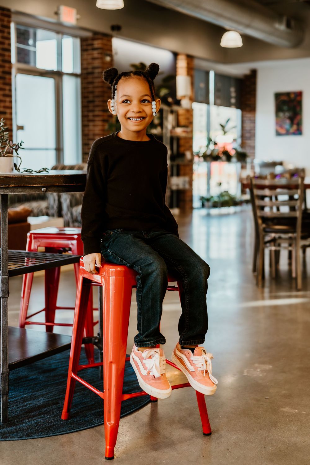 Young person in black sweater and jeans sits on red metal stool in modern industrial cafe space.