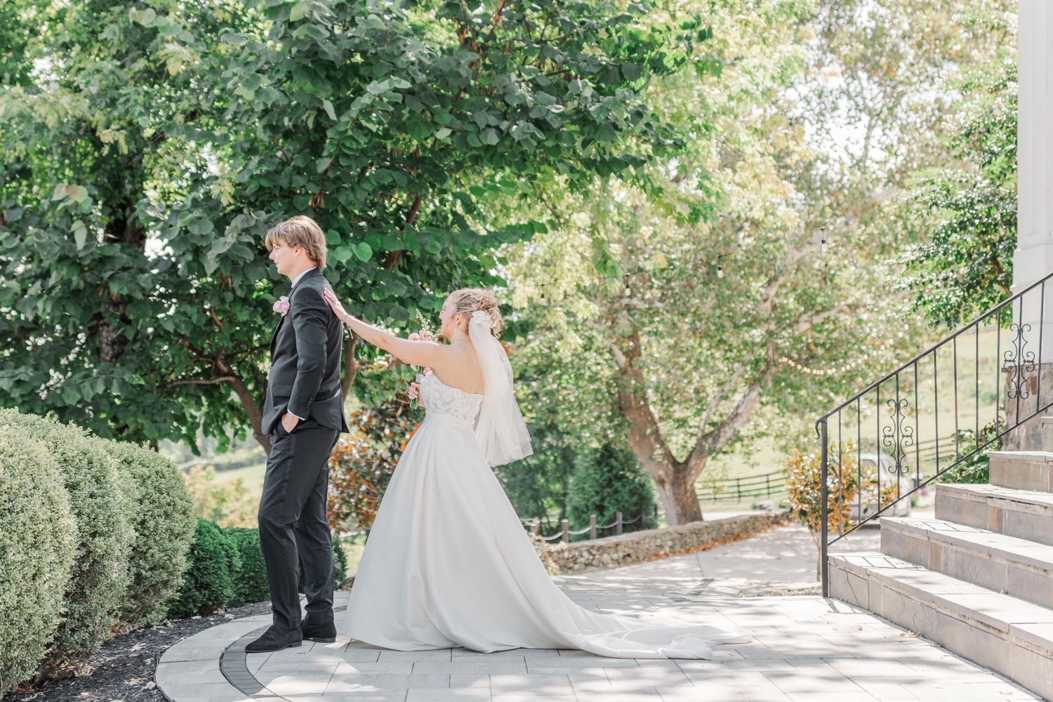 Newlyweds walk along garden path with flowing wedding dress train in natural outdoor setting.