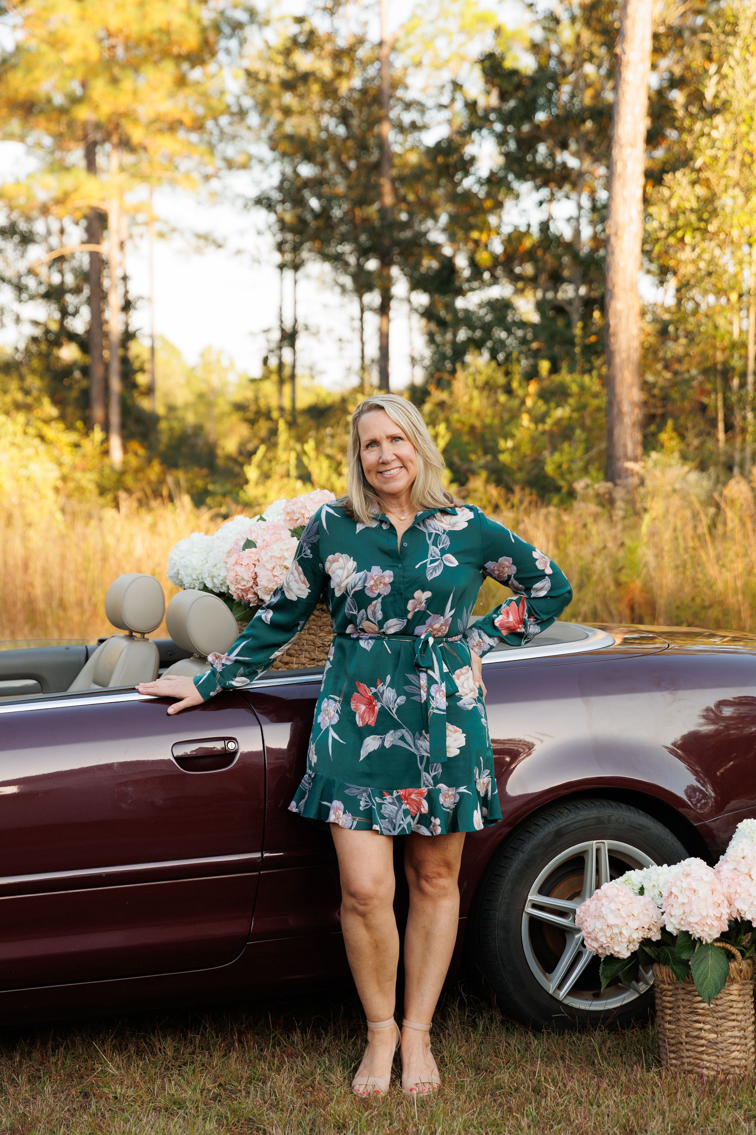 A person in a floral dress poses next to a burgundy convertible car surrounded by autumn trees and pink flowers.