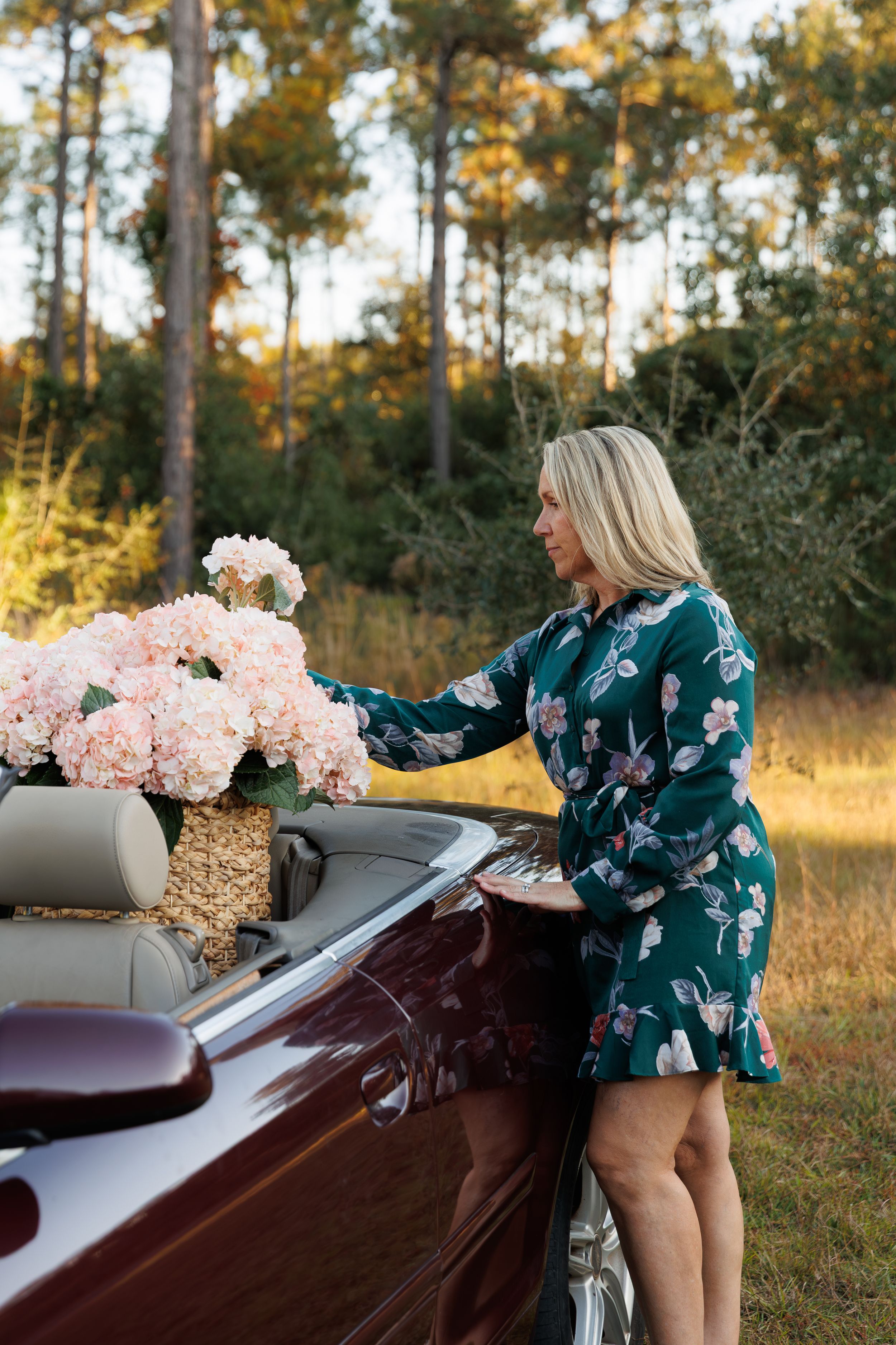 A person in a floral green dress arranges pink peonies on a vintage car in a sunlit outdoor setting.