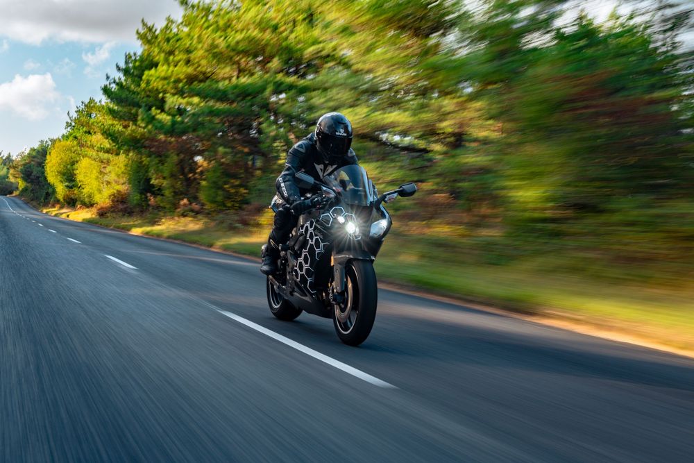 A motorcyclist rides a black sports bike at high speed on a curved road with motion blur and trees in background.