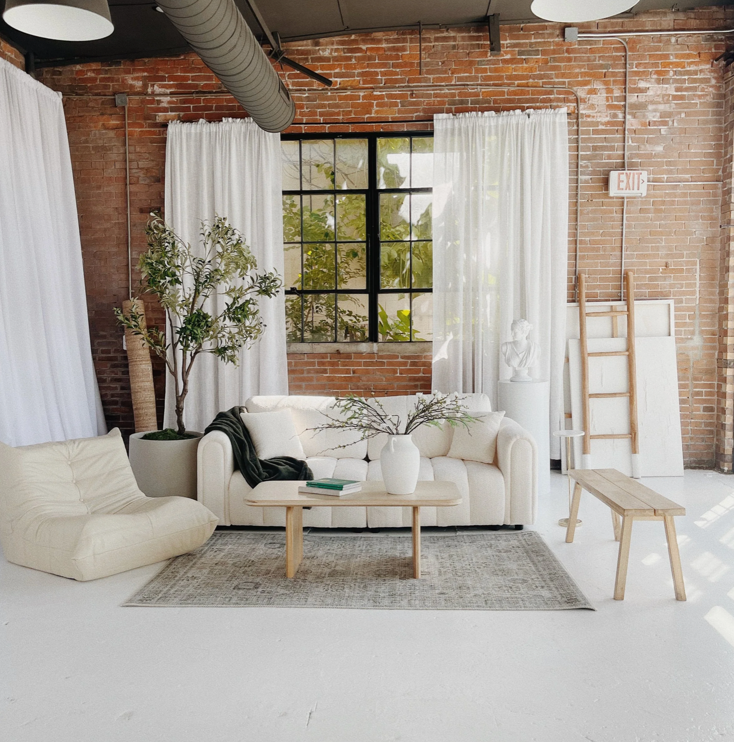 Modern industrial loft space with exposed brick walls, white curtains, and minimalist furniture in a bright open layout.