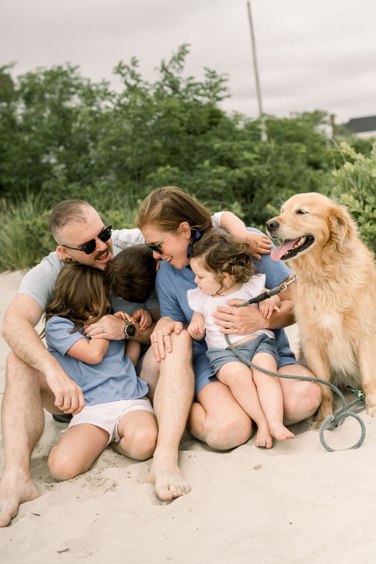 A golden retriever dog lies on a sandy beach with people in casual summer clothing relaxing together.