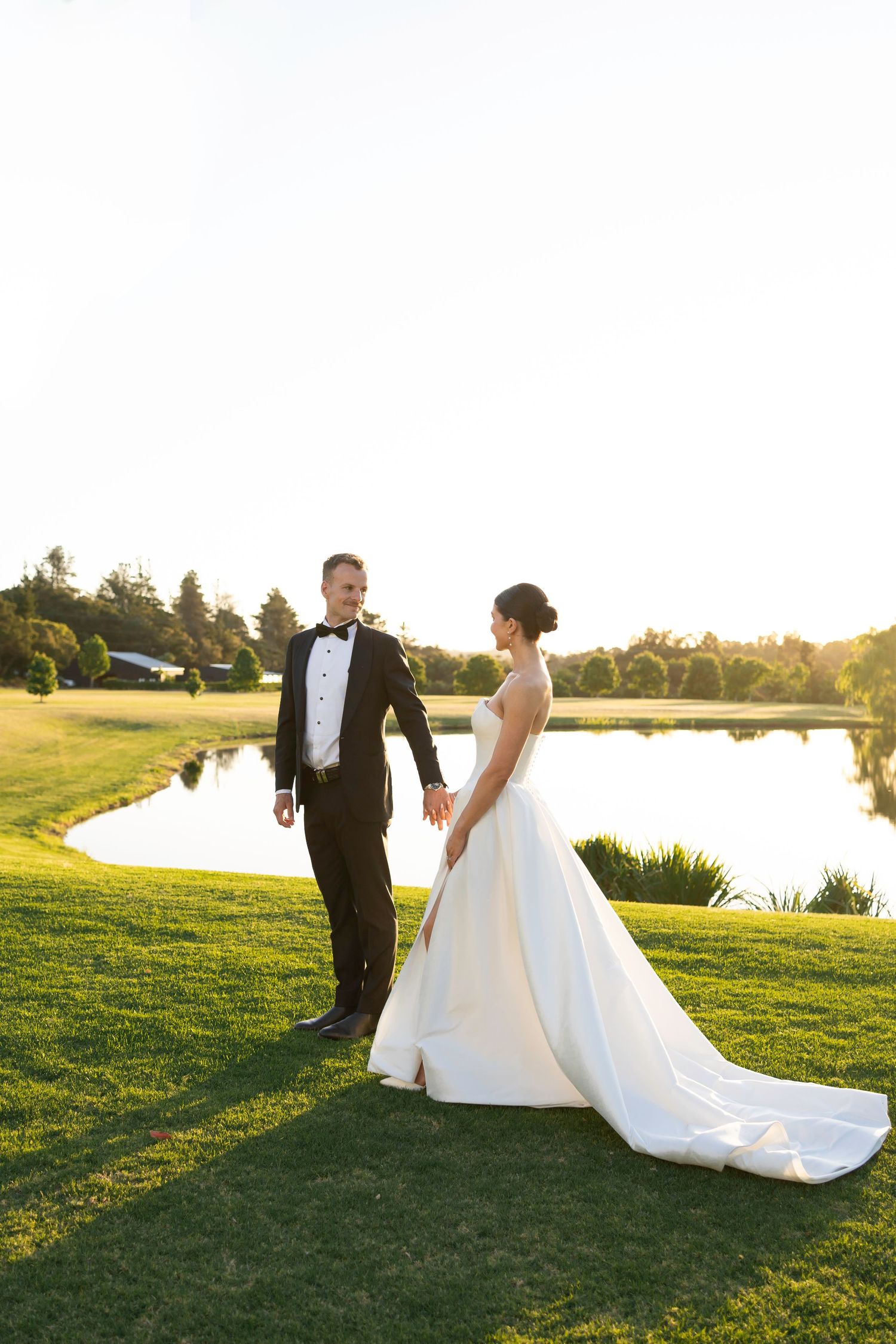 Newlywed couple walks together on a scenic golf course during golden hour sunset by a pond.