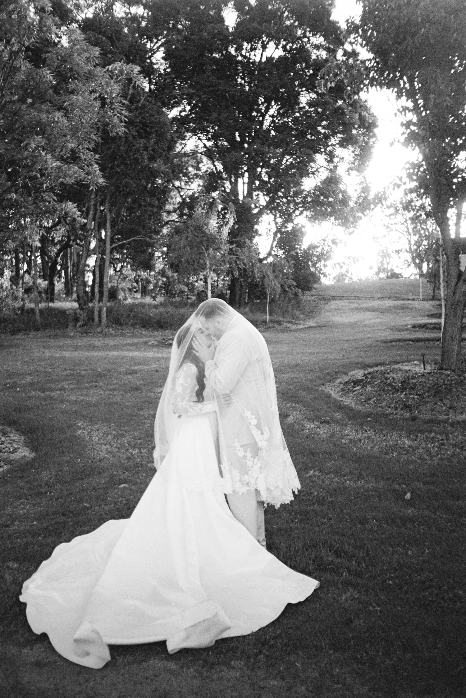A bride in a flowing white wedding dress and veil stands on a tree-lined path in a black and white photograph.