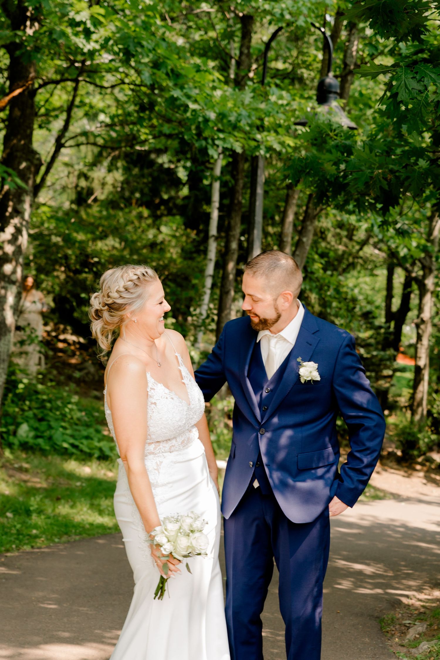 Bride and groom share a joyful moment on a tree-lined path, she holds a white bouquet in a lace gown.