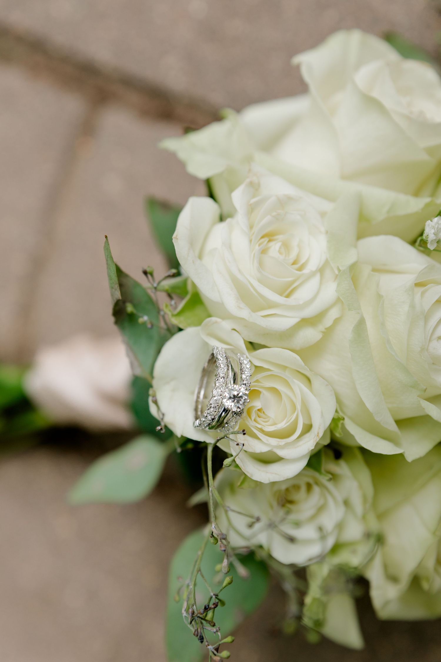 Wedding rings nestled among white roses in a bridal bouquet on a stone surface.