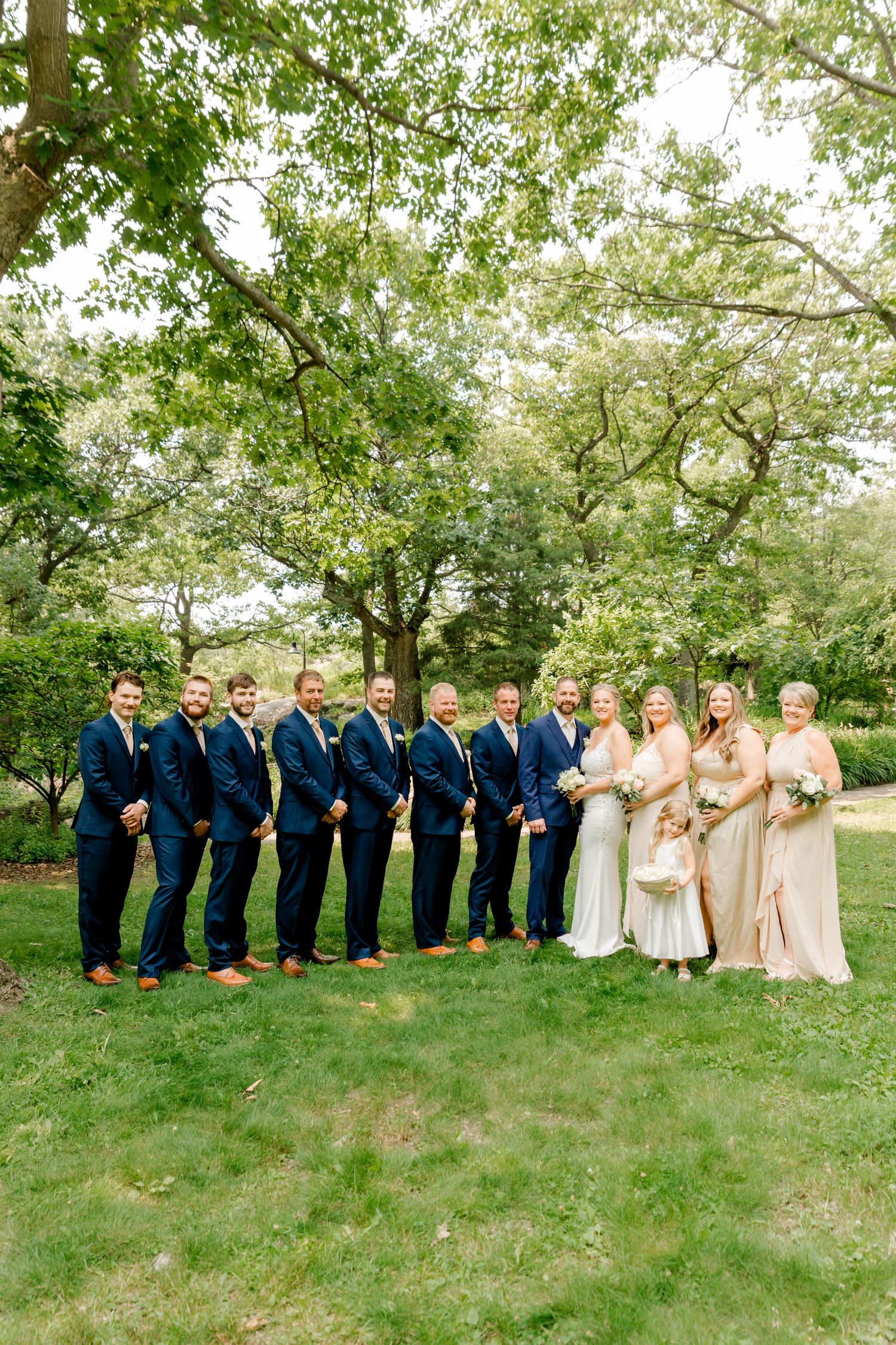 Wedding party poses outdoors, groomsmen in navy suits and bridesmaids in champagne gowns under a large tree.
