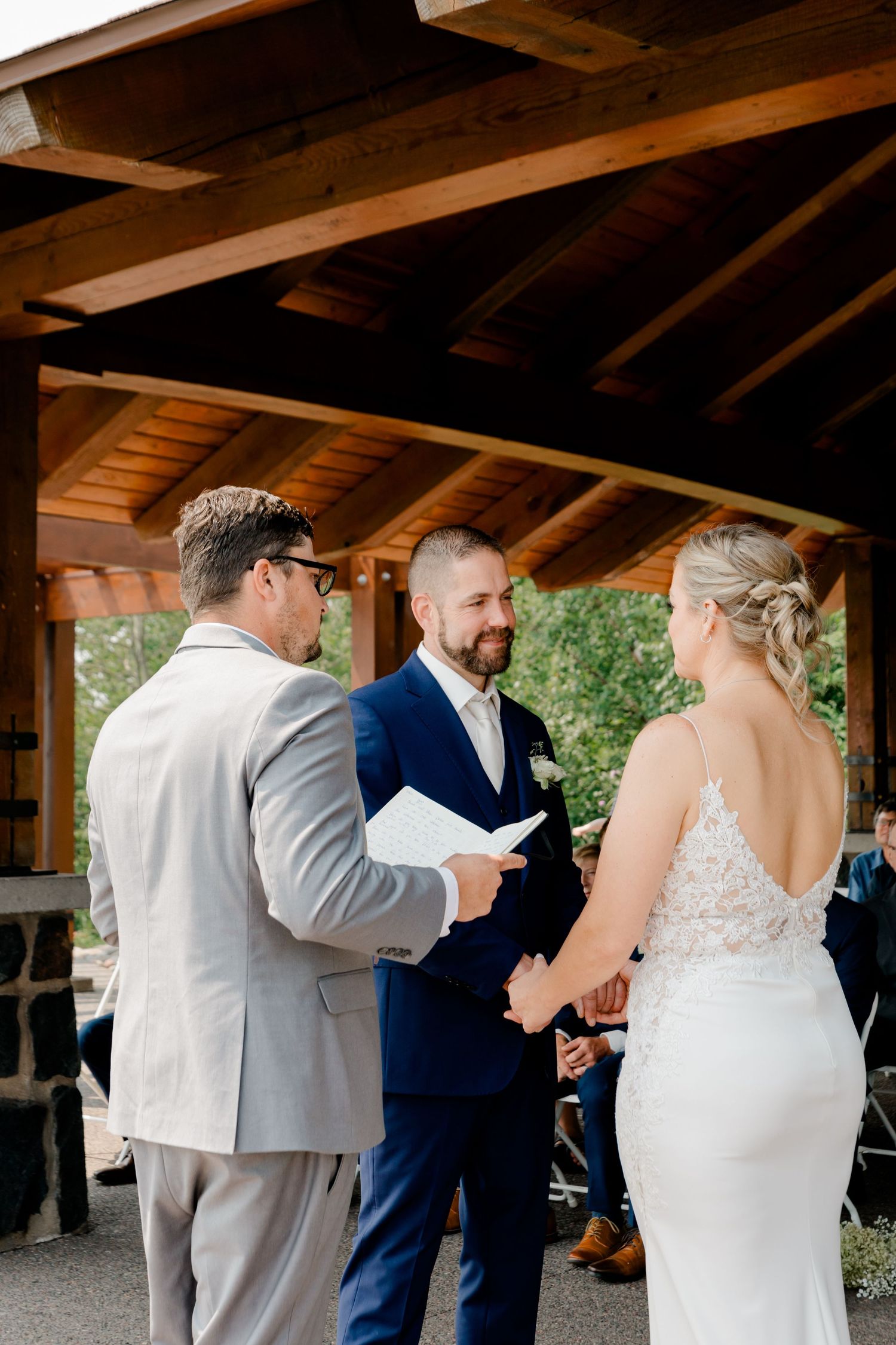 Groom smiles during outdoor wedding ceremony under wooden pavilion as officiant reads vows to couple.