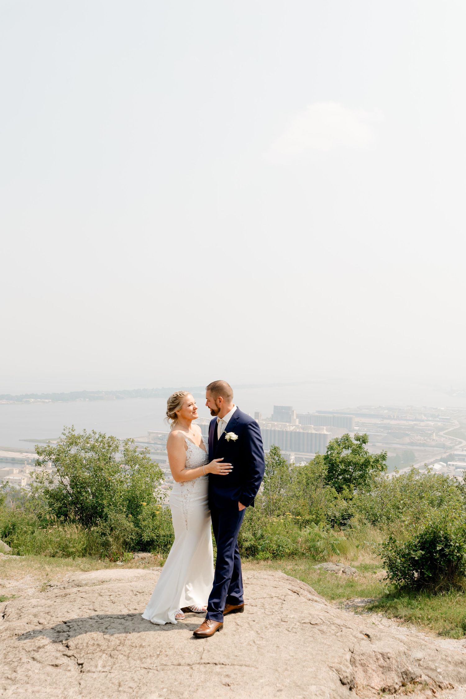 Bride and groom stand on rocky overlook with scenic waterfront cityscape behind them on wedding day.
