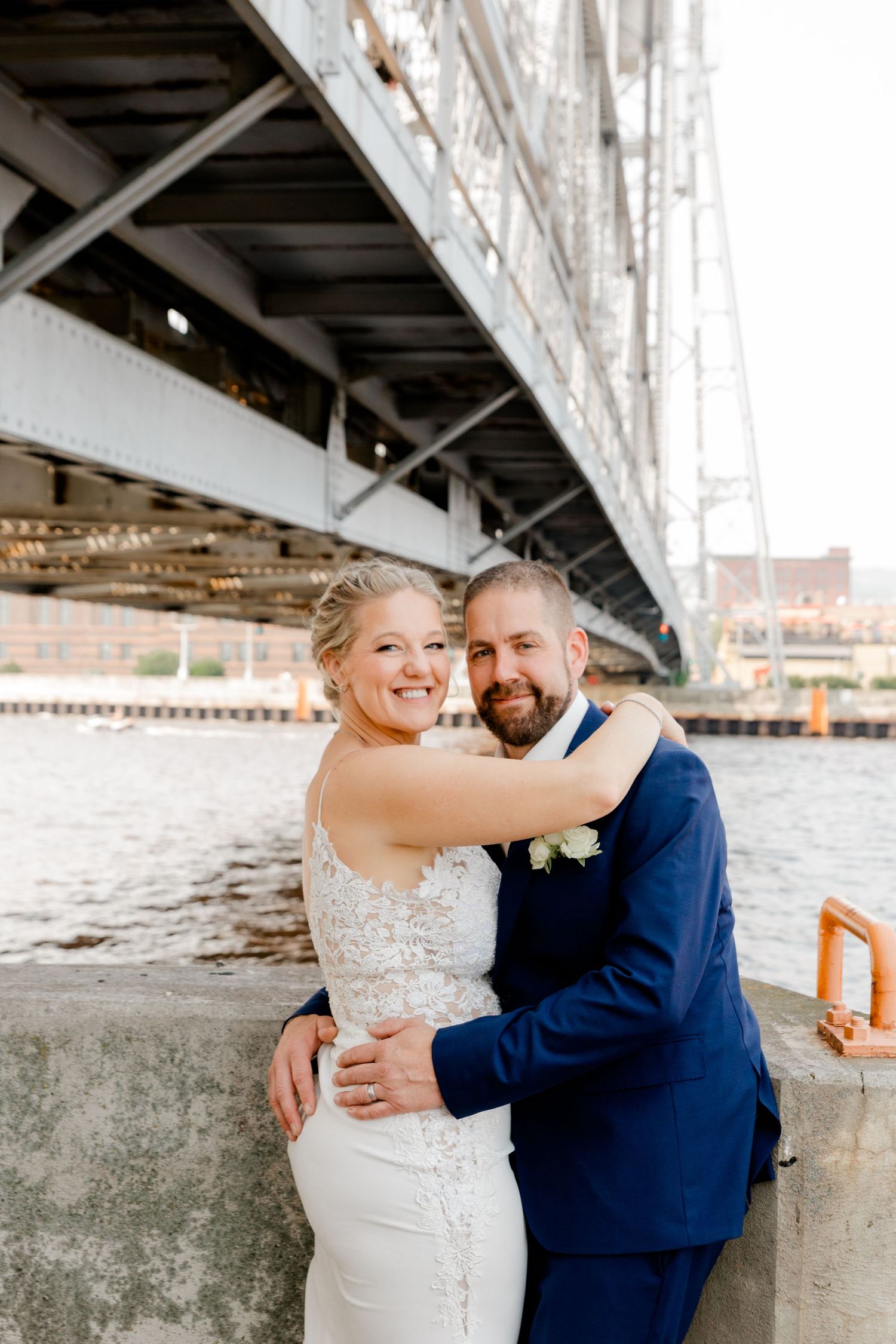 Smiling bride and groom embrace by the waterfront under a large steel bridge on their wedding day.