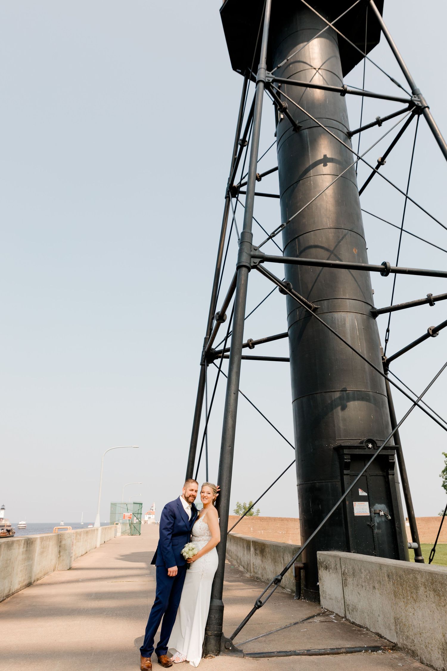Bride and groom pose together beside a tall black industrial tower structure on a sunny wedding day.