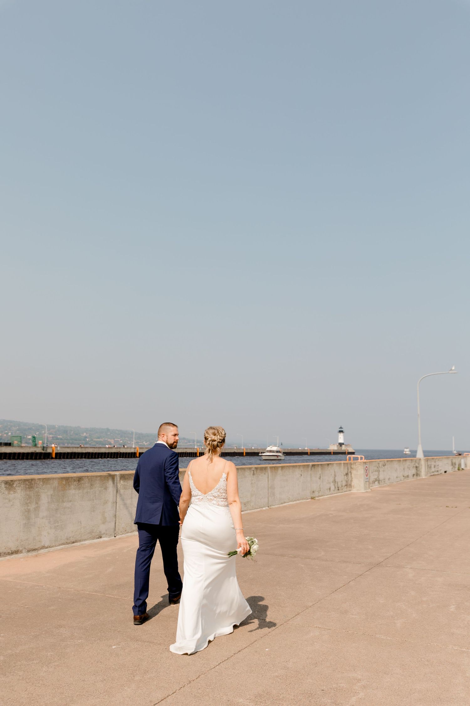 Bride and groom walk hand in hand along a waterfront pier with a lighthouse in the distance.