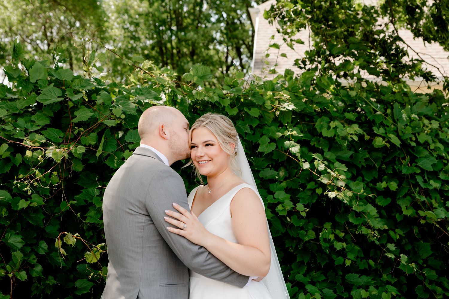 Groom whispering to smiling bride surrounded by lush green foliage in garden portrait.