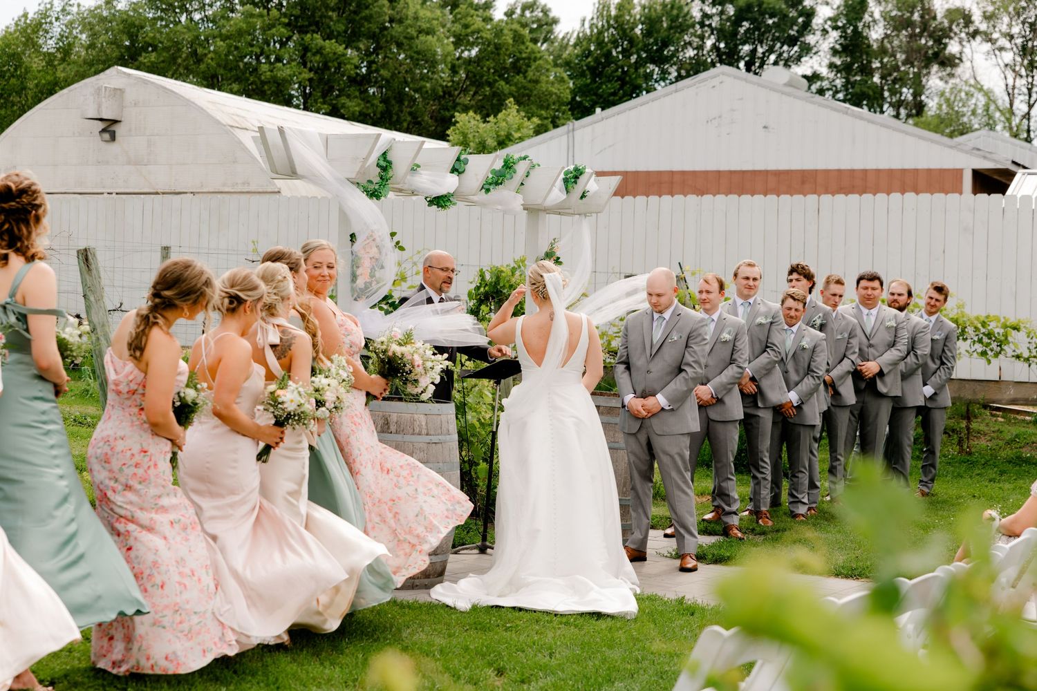 Wedding party gathered outdoors on green lawn, bridesmaids in pink and sage dresses facing groomsmen in gray suits.