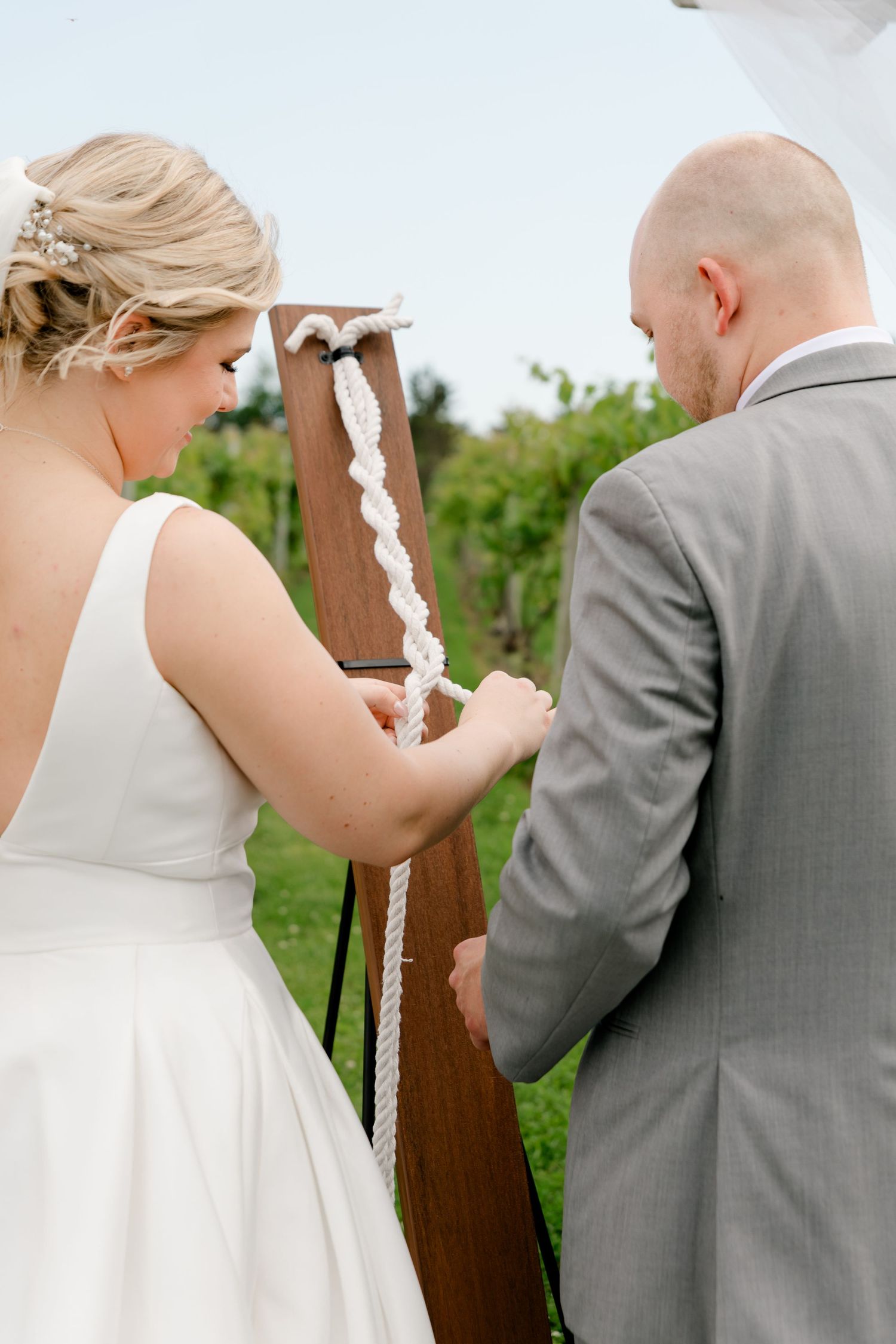 Bride and groom performing unity rope ceremony at wooden cross during outdoor vineyard wedding.