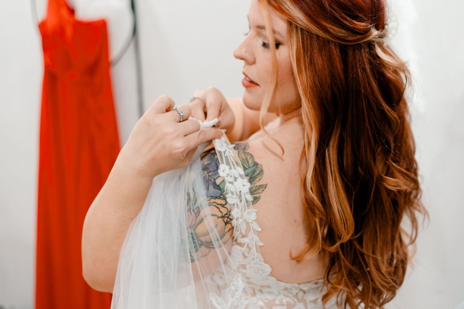Red-haired bride with floral tattoo adjusting veil on lace wedding dress in bright dressing room.