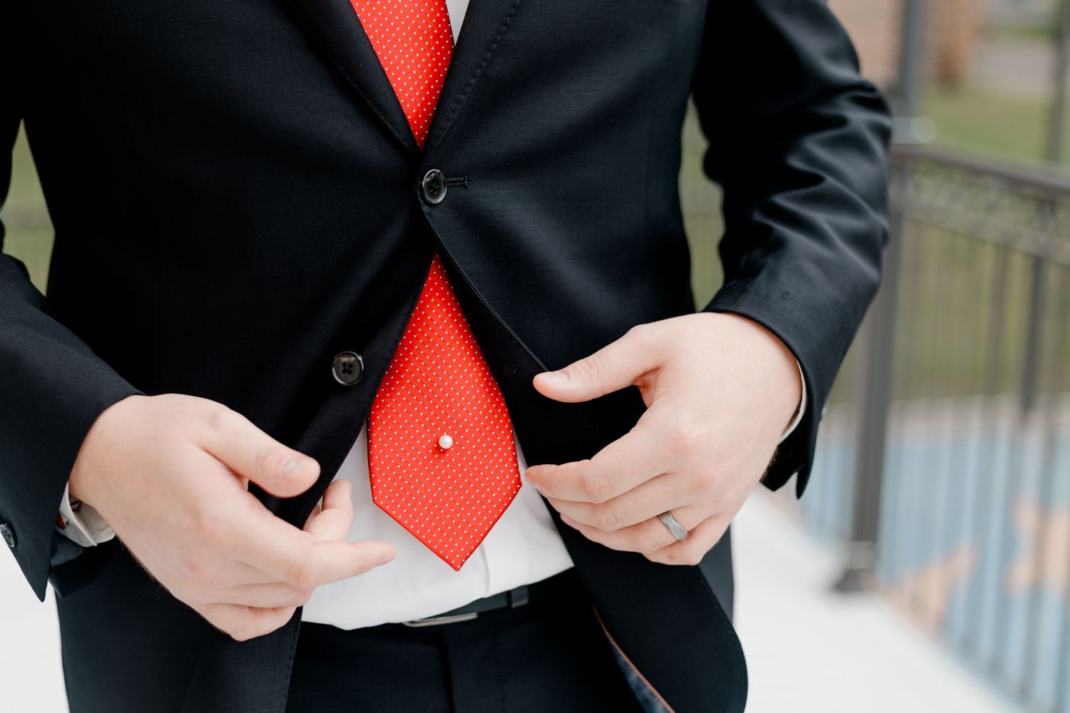 Close-up of groom in black suit adjusting jacket with red tie and boutonniere detail.