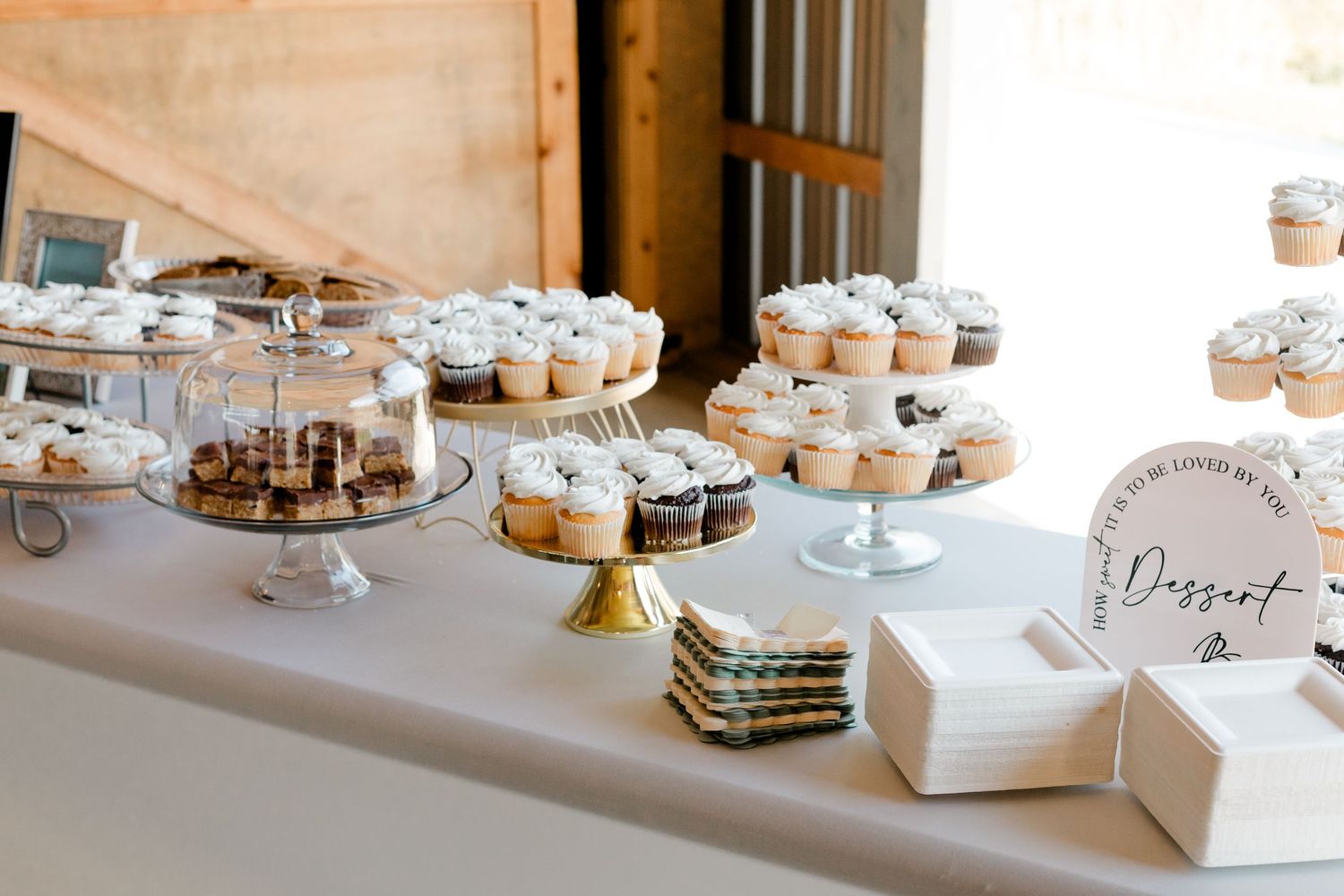 Elegant wedding dessert table with white frosted cupcakes on tiered stands and a decorative dessert sign in a rustic barn.