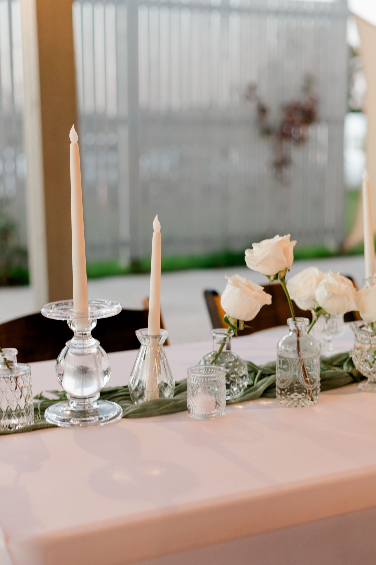 Elegant wedding table with white roses, tapered candles on crystal holders, and small glass vases on a blush tablecloth.