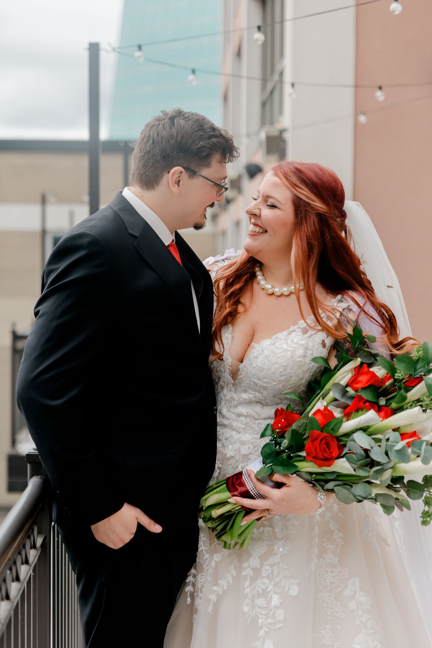 Smiling bride with red hair holding red rose bouquet stands close with groom in black tuxedo outdoors.