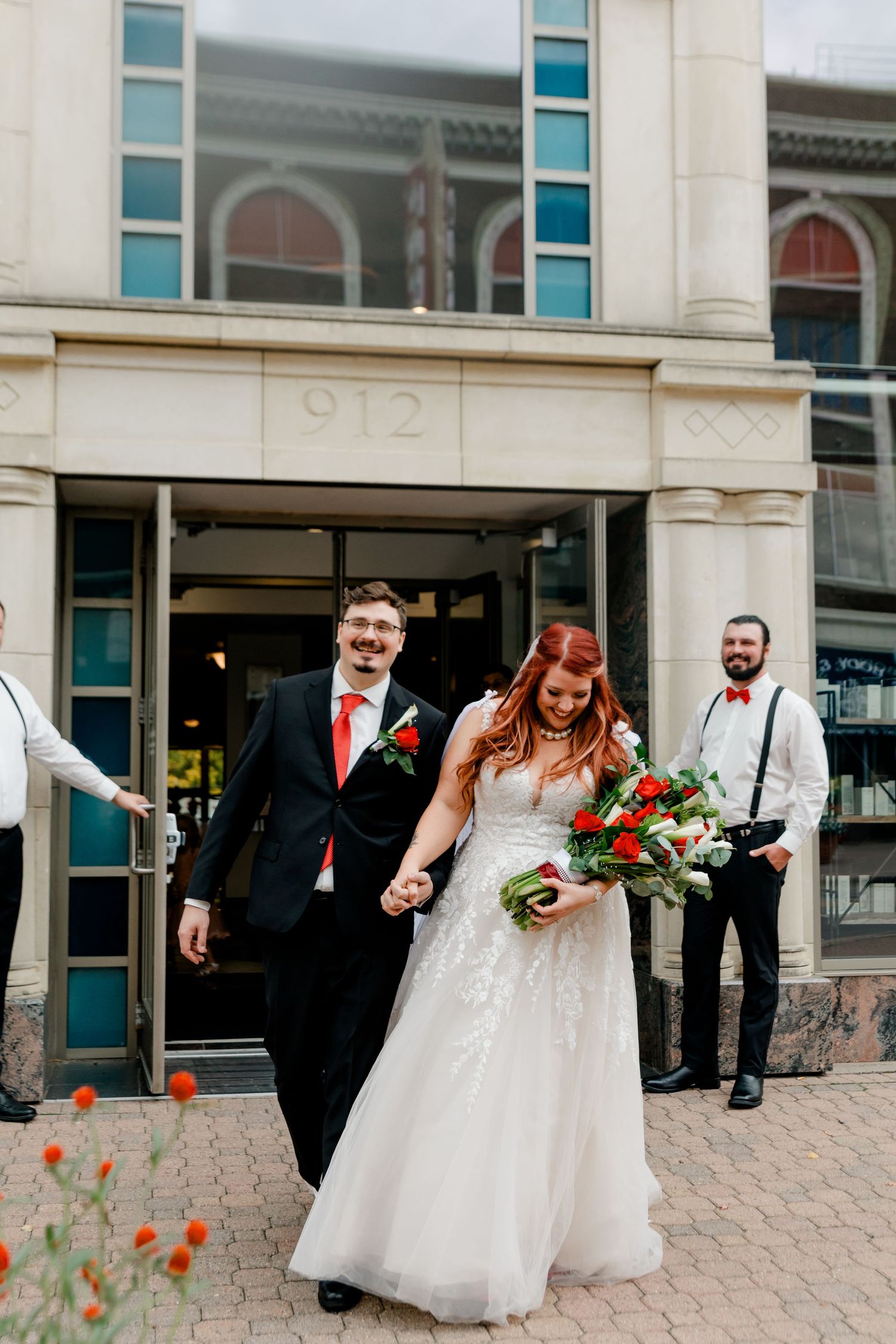 Newlyweds exit building 912 hand in hand, bride in lace gown holding red floral bouquet, groom in black suit.