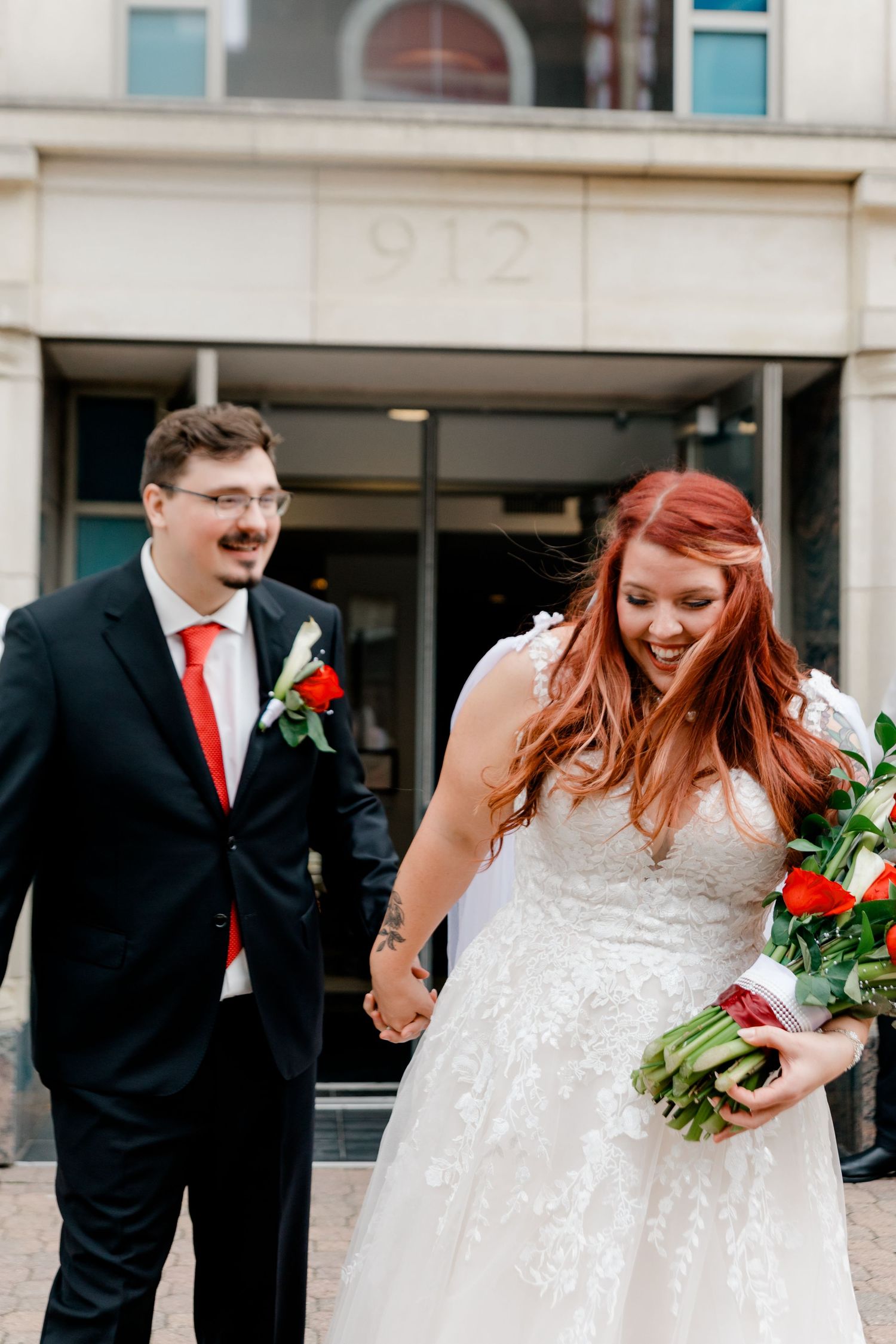 Laughing bride with flowing red hair and groom in black suit walk hand in hand outside building 912.