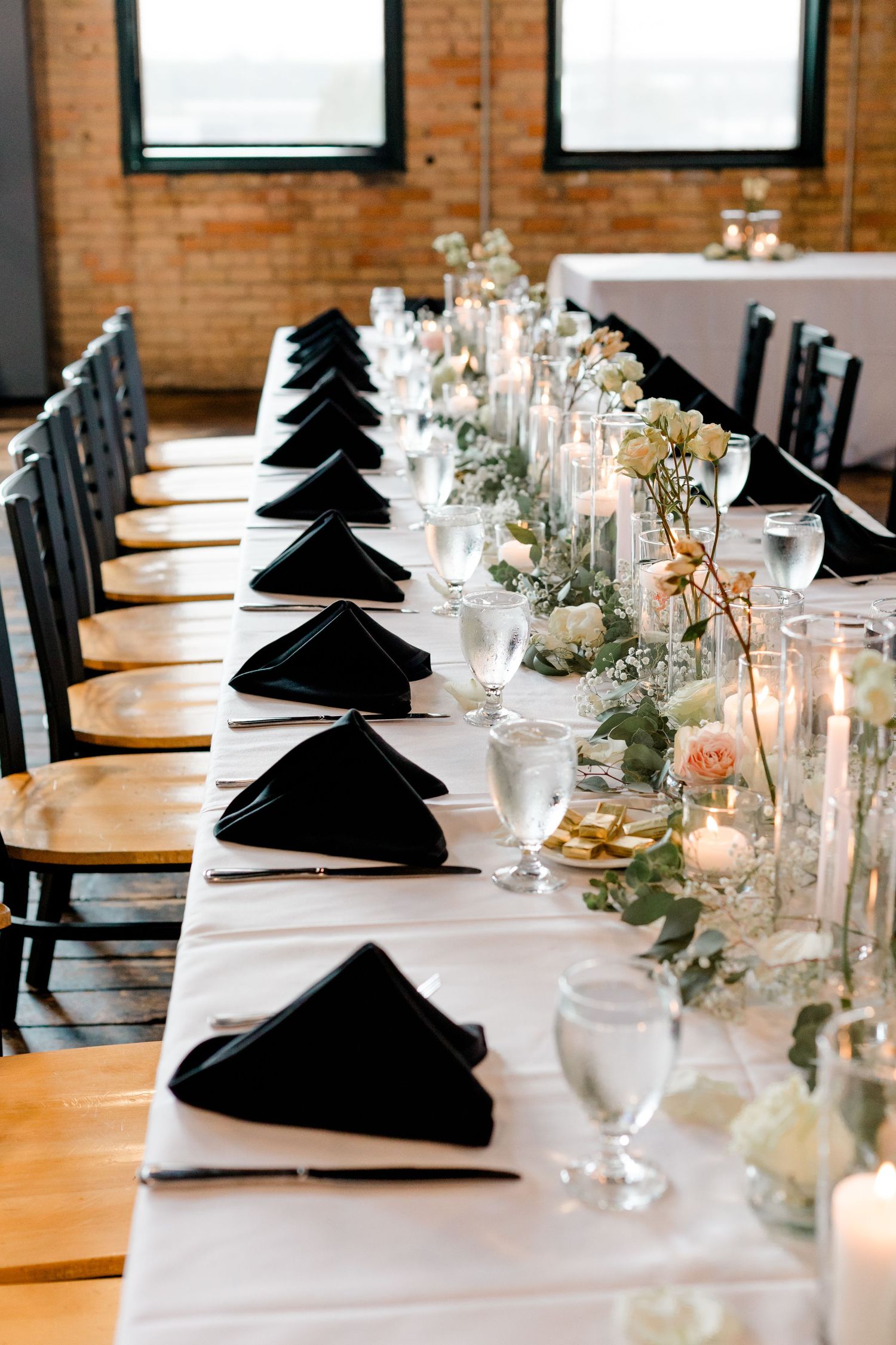 Elegant wedding reception table with black napkins, white linens, floral centerpieces, and candles in a brick venue.