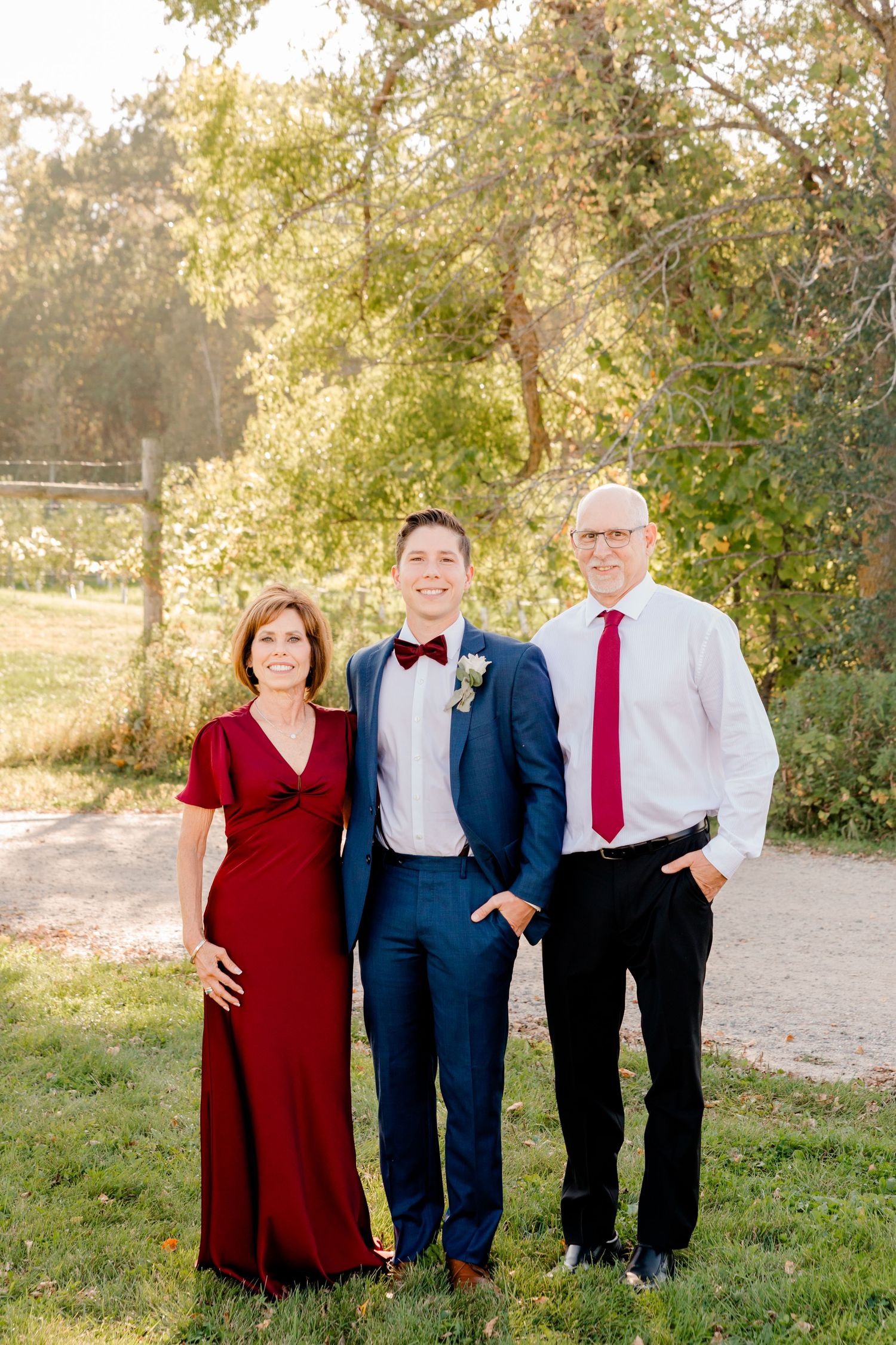 Groom in navy suit poses with parents in burgundy formal attire outdoors at golden hour.