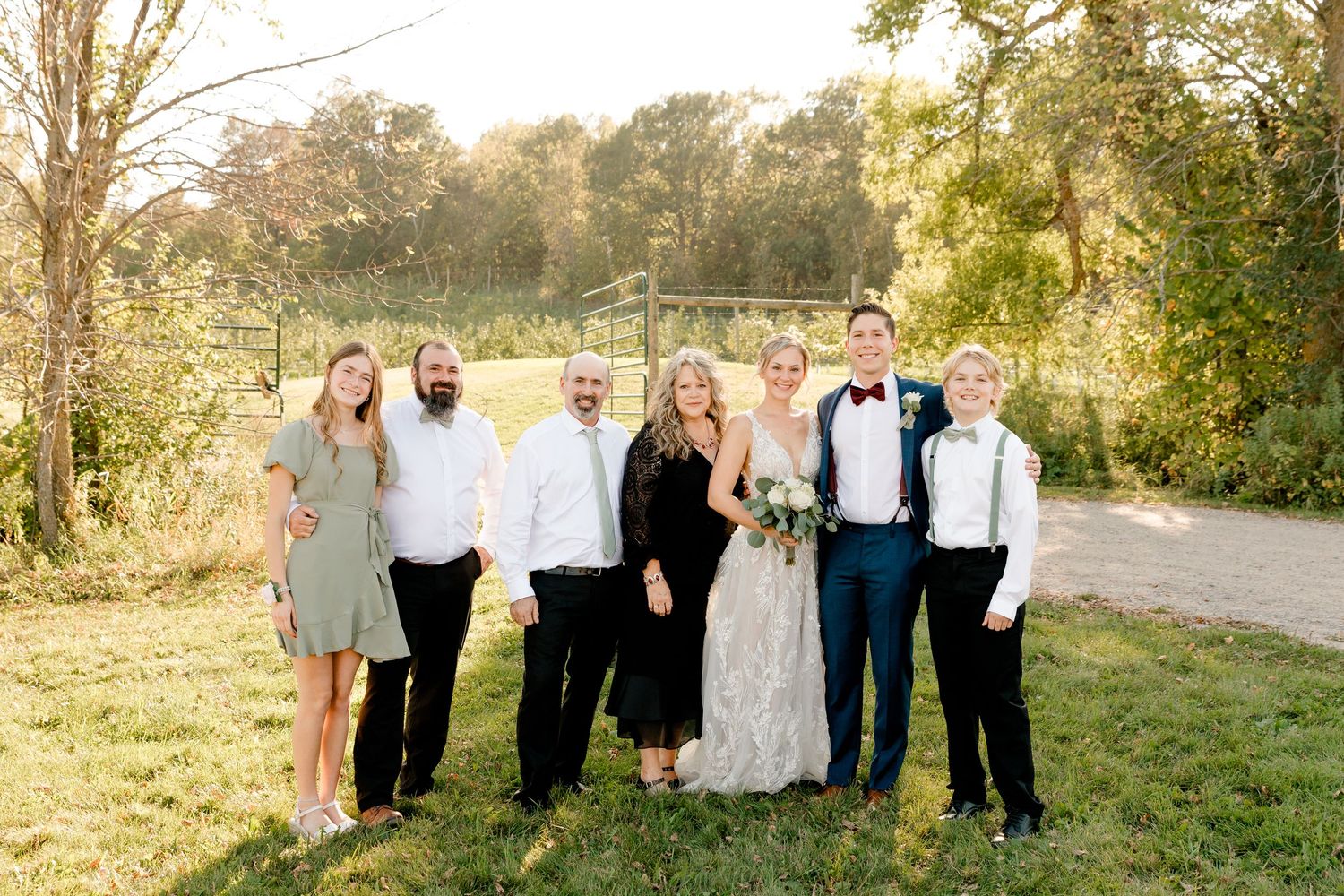 Wedding party group portrait of bride, groom, and family members smiling outdoors at sunset.