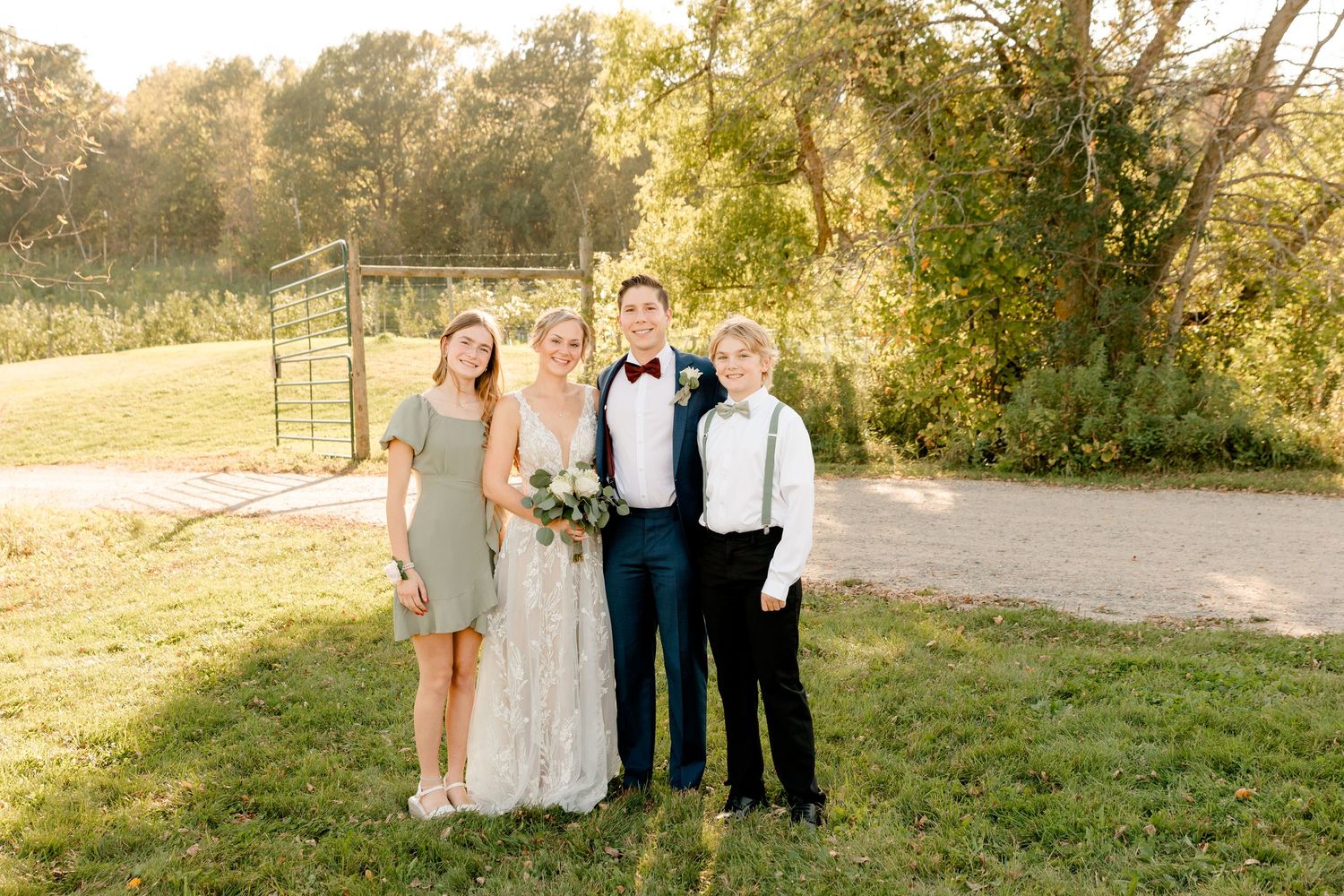 Bride and groom pose with two children on a sunny lawn at an outdoor wedding venue surrounded by lush green trees.