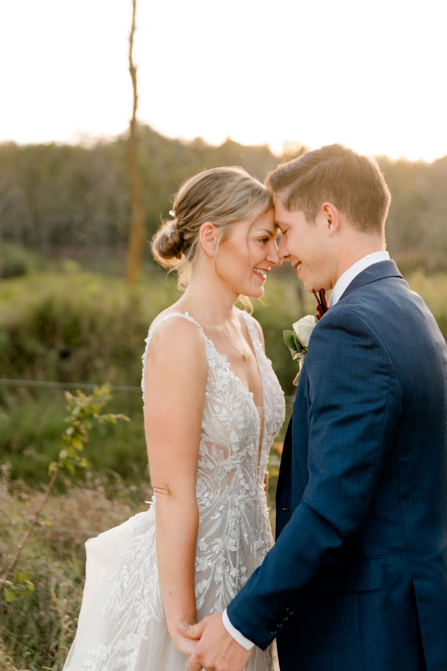 Bride and groom touch foreheads while holding hands outdoors at golden hour, she in lace gown, he in navy suit.