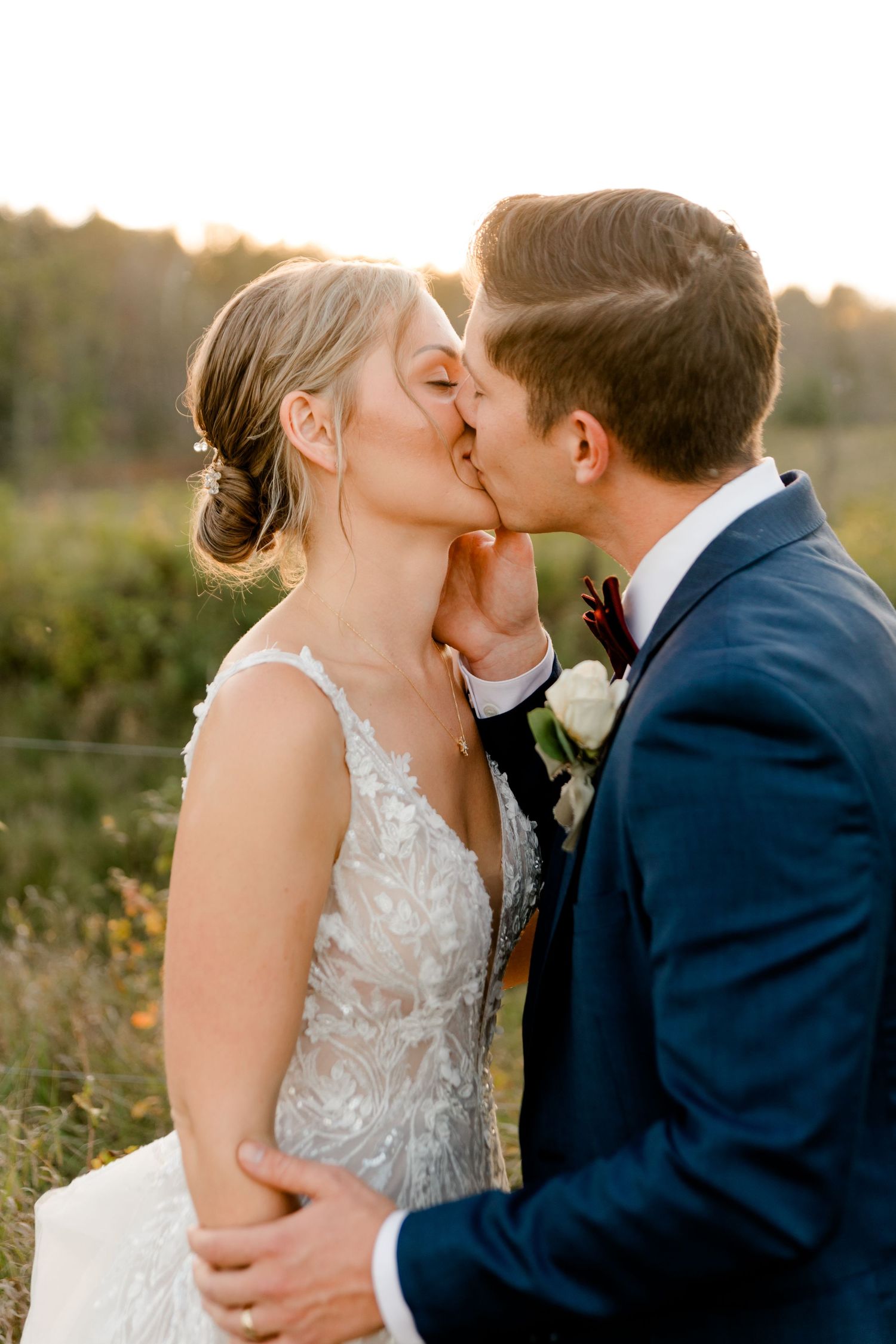 Bride and groom share a romantic sunset kiss outdoors, wearing wedding dress and navy suit with floral boutonniere.