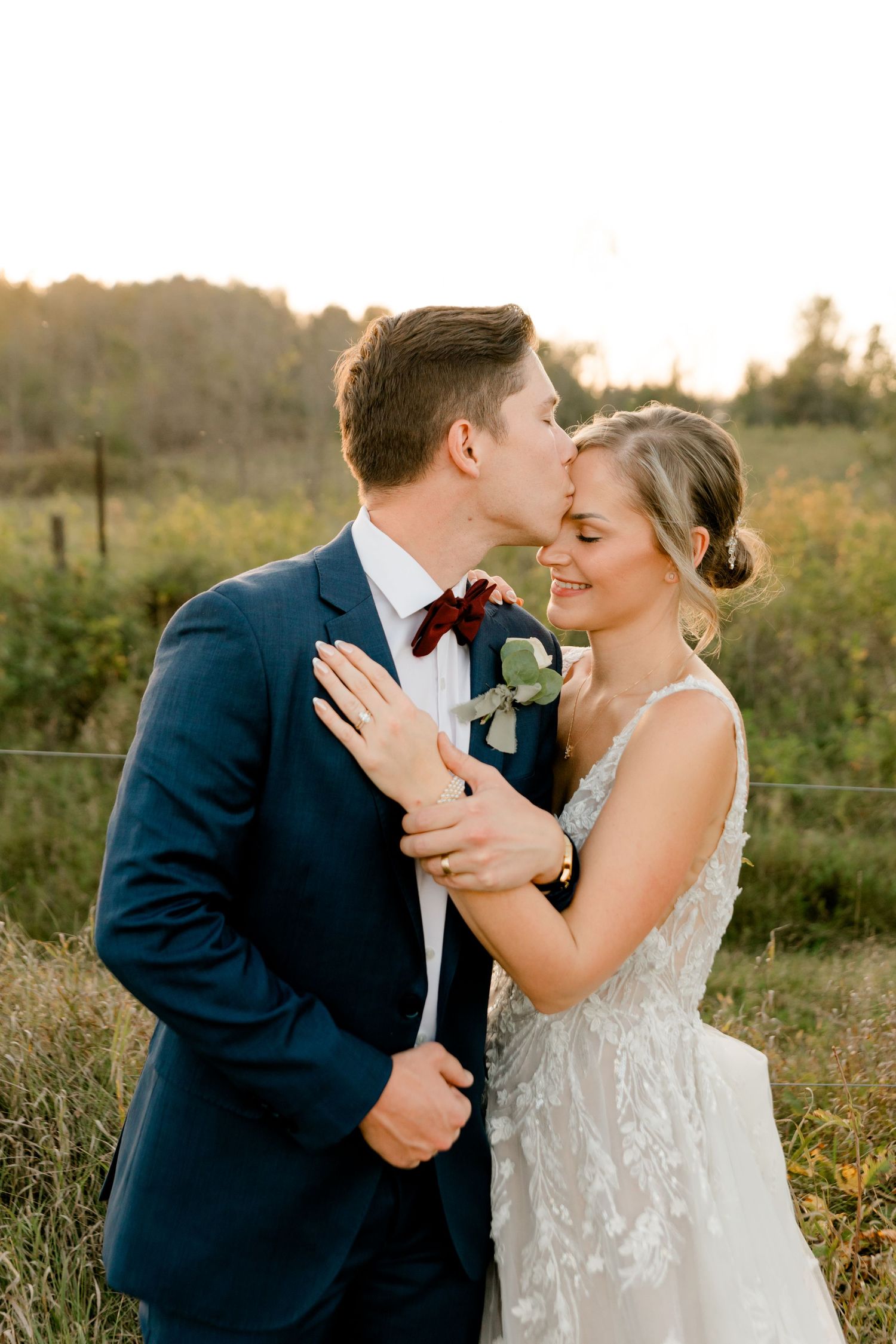 Bride and groom share a tender moment at golden hour, groom kissing bride's cheek in a lace gown and navy suit.