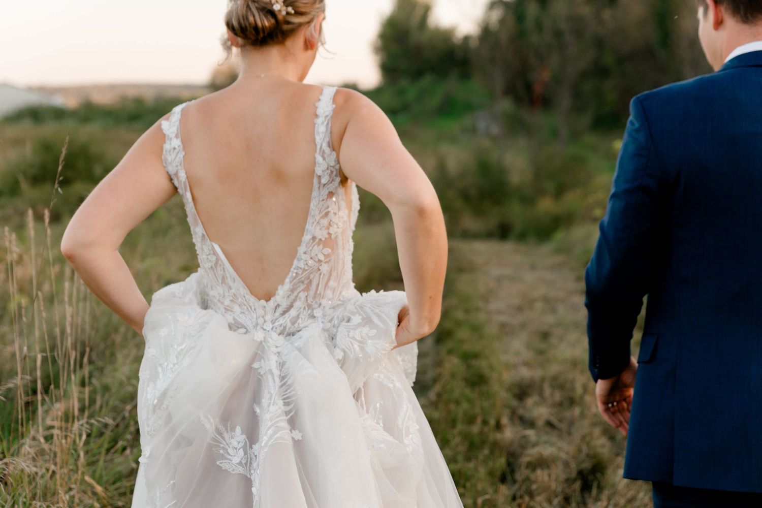 Bride in elegant backless lace wedding gown walks hand-in-hand with groom in navy suit through scenic outdoor field.