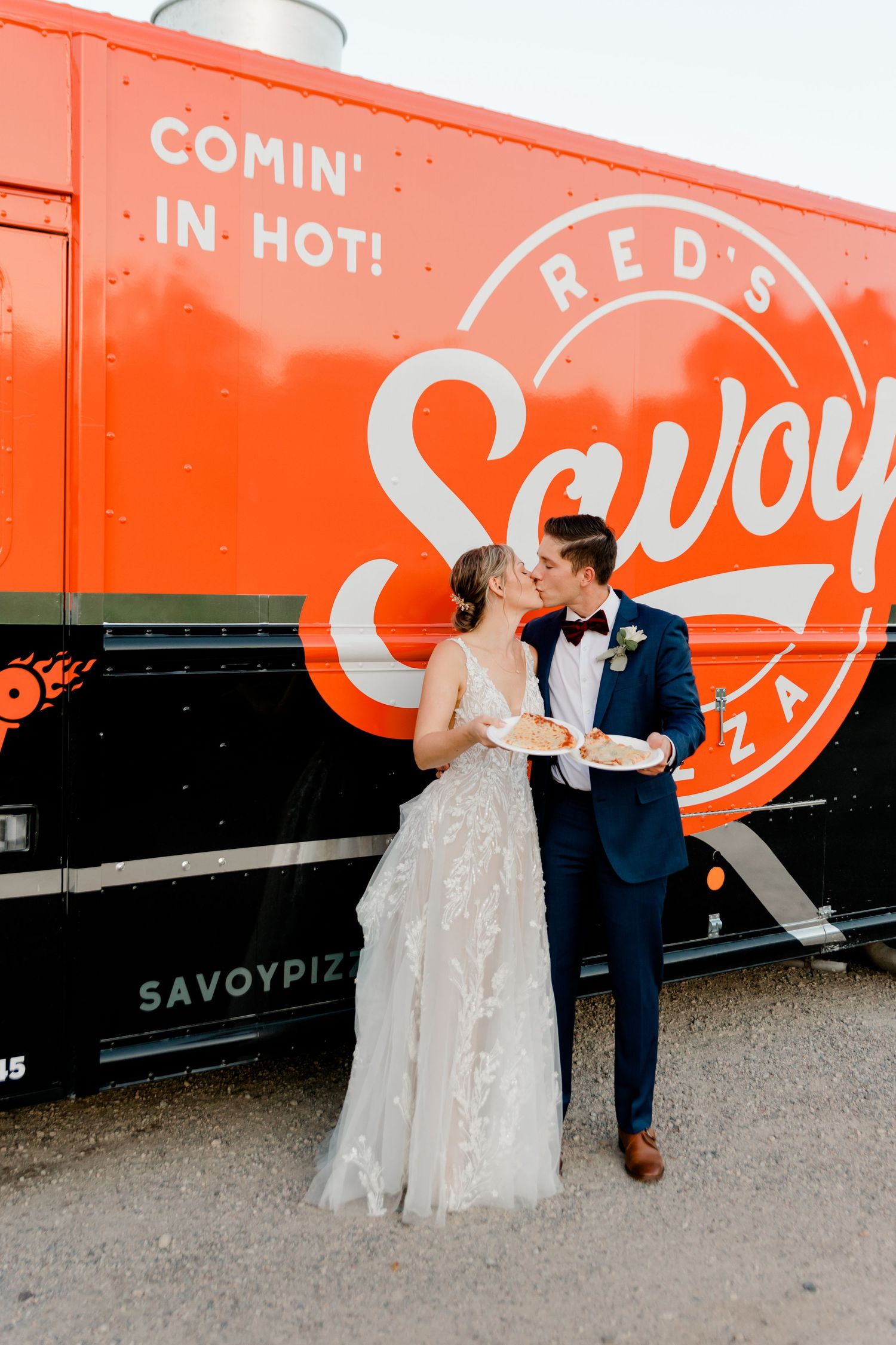 Newlyweds share a kiss while holding pizza slices in front of Red's Savoy Pizza food truck at their wedding.