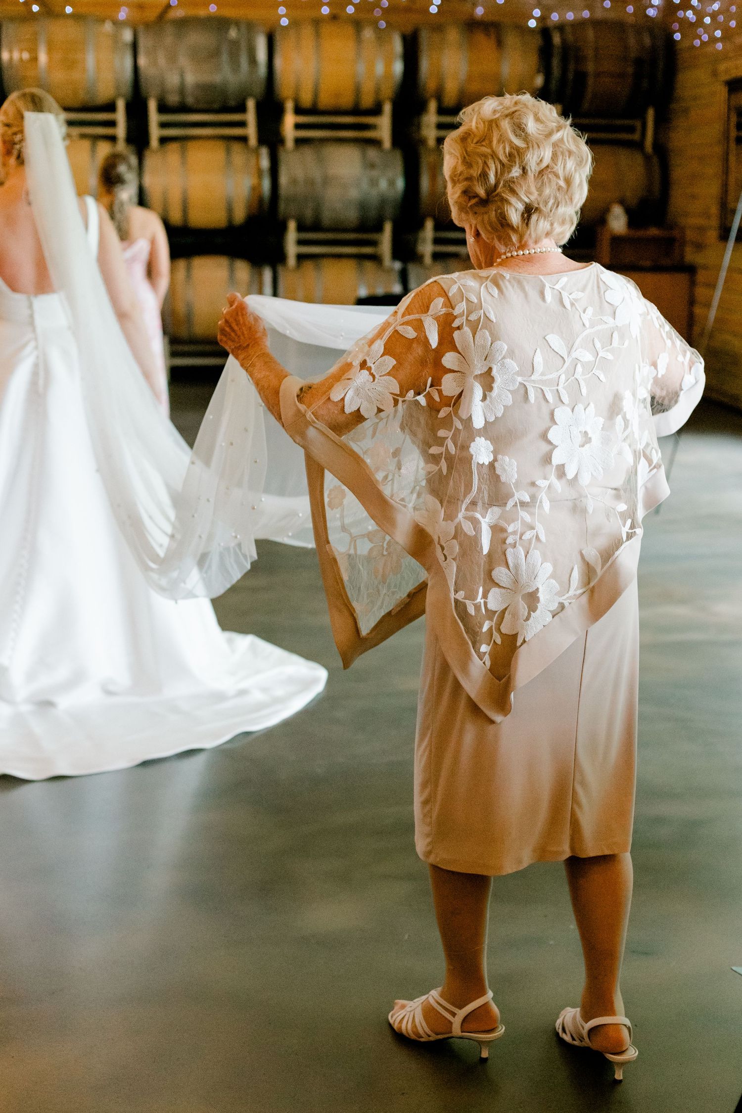 Older woman in floral applique dress helping bride with veil in winery barrel room.