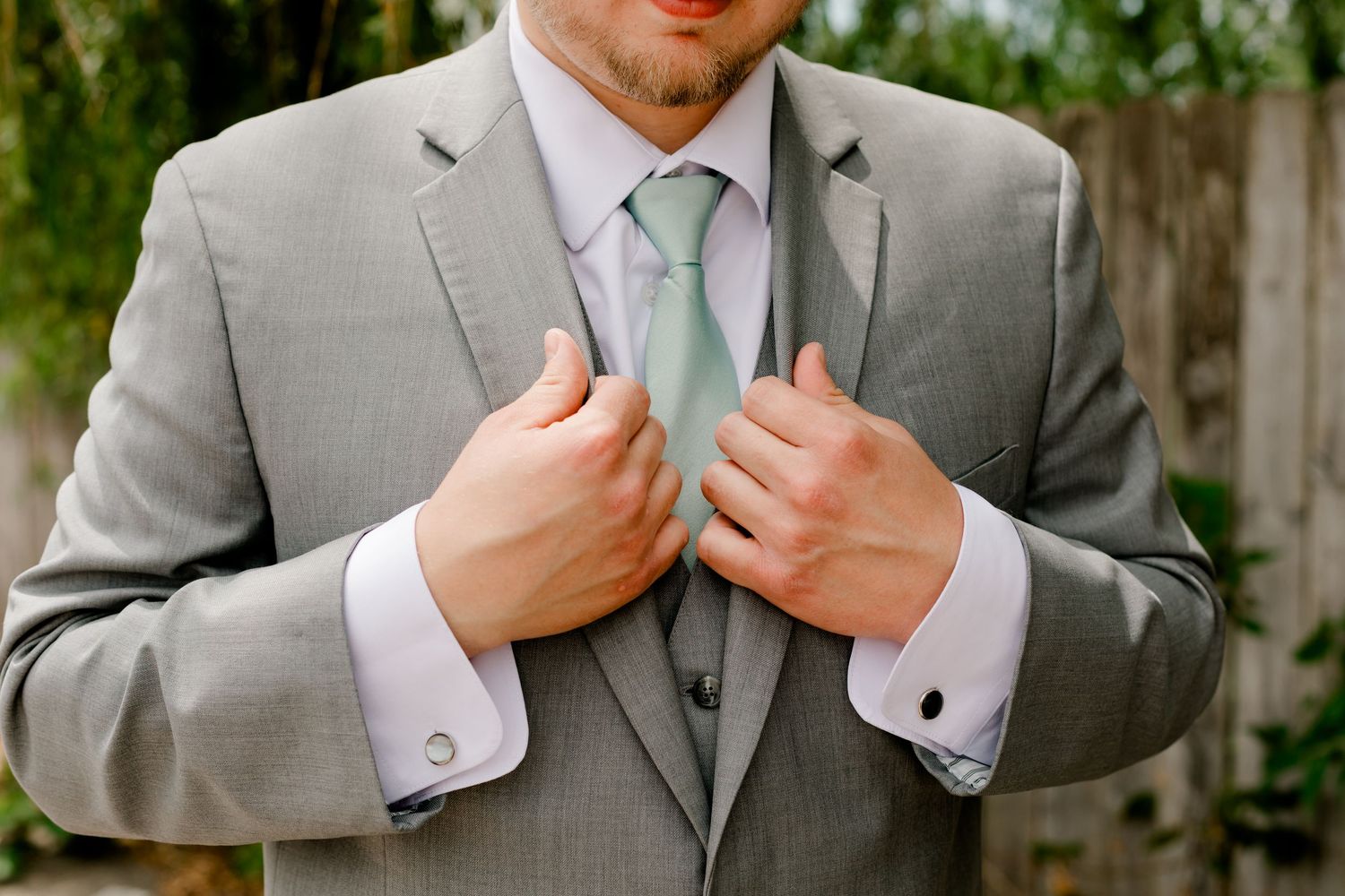 Groom in grey suit adjusting mint green tie with cufflinks before wedding ceremony.