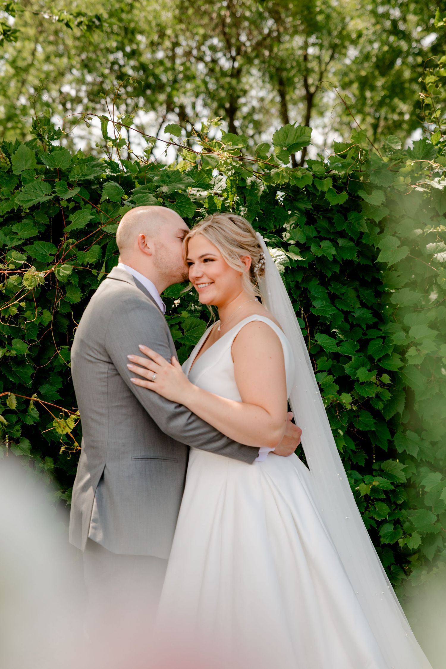 Groom kissing bride on cheek as she smiles in lush green garden setting.