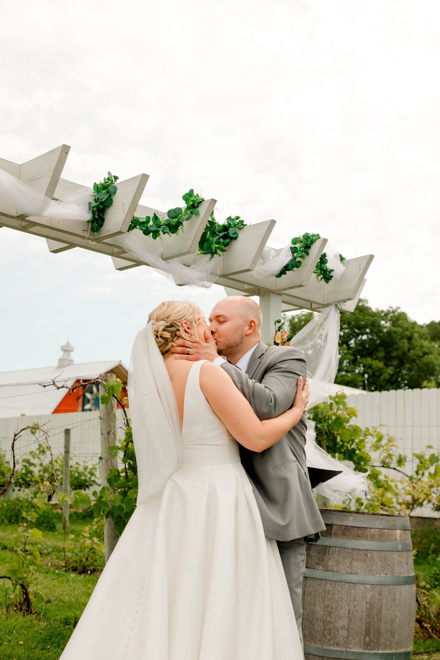 Bride and groom sharing first kiss under white pergola decorated with greenery at vineyard.