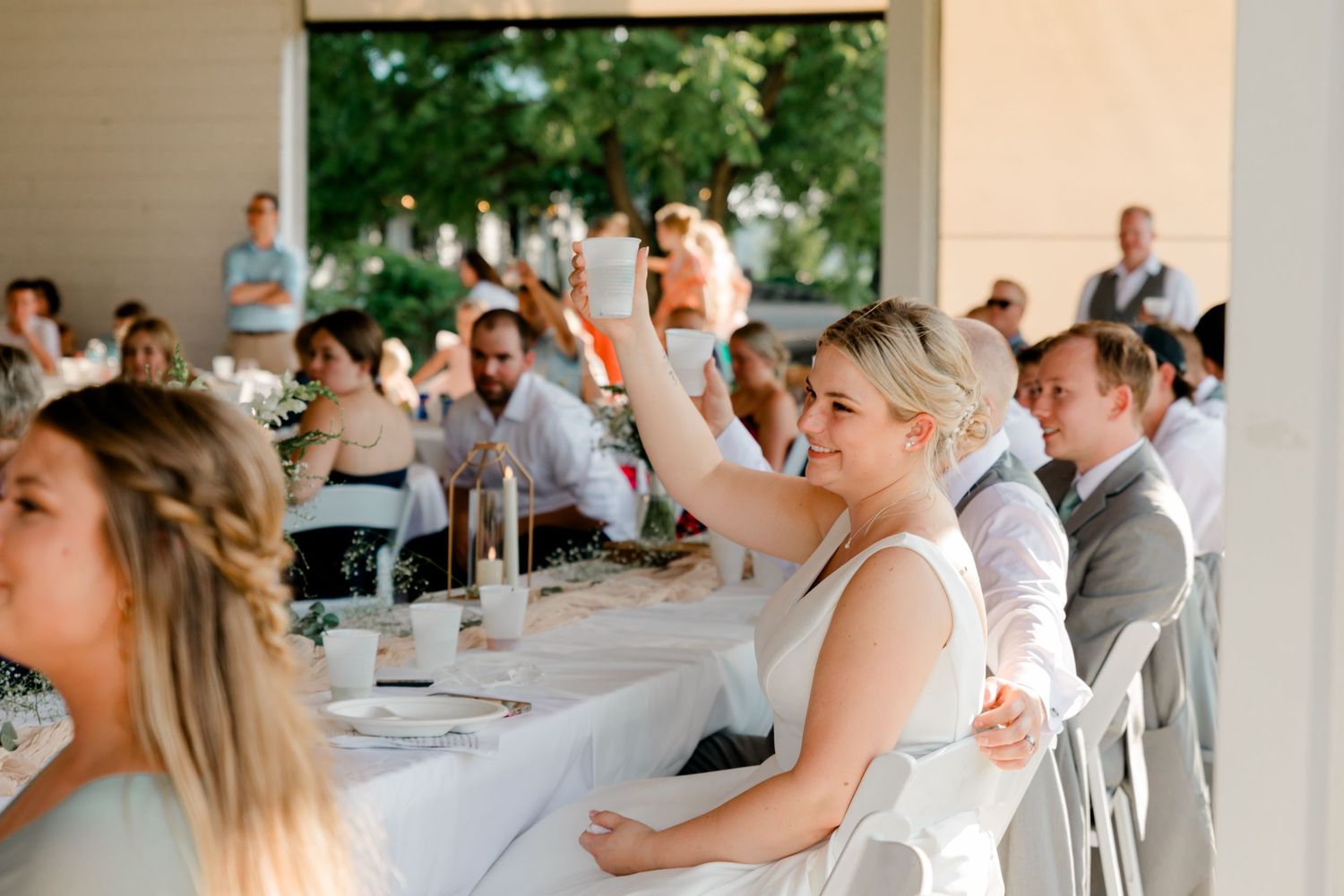 Smiling bride raising hand at outdoor wedding reception dinner with guests at long table.