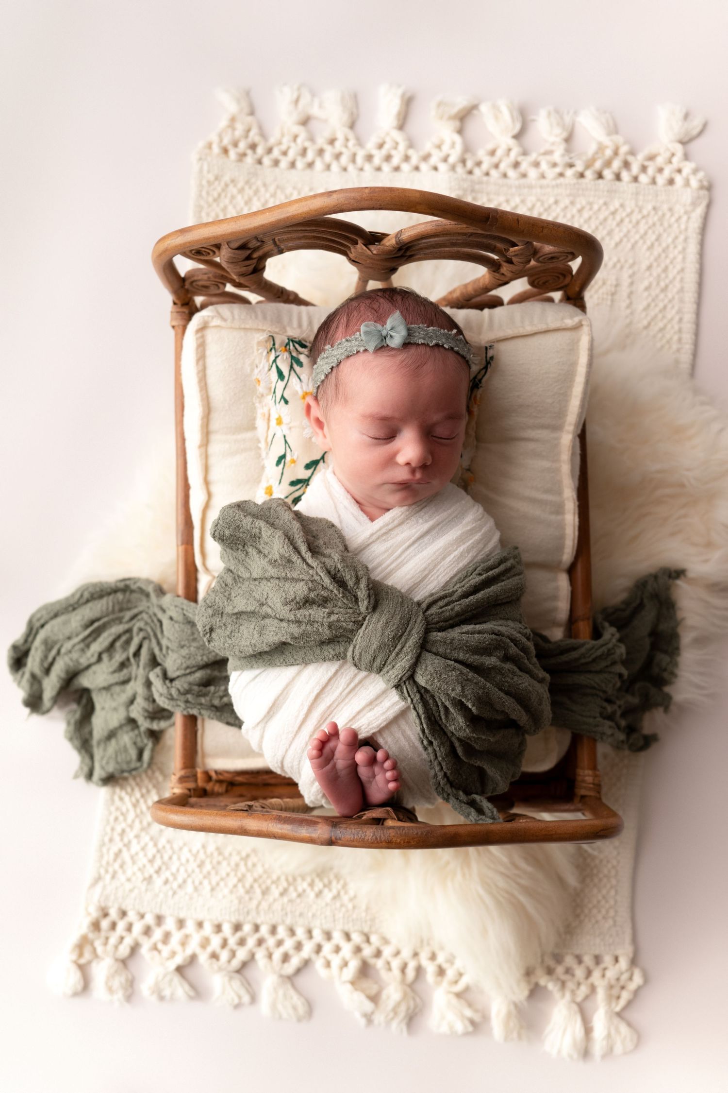 A sleeping newborn baby wrapped in sage green fabric lies peacefully in a rustic wooden basket on white lace bedding.